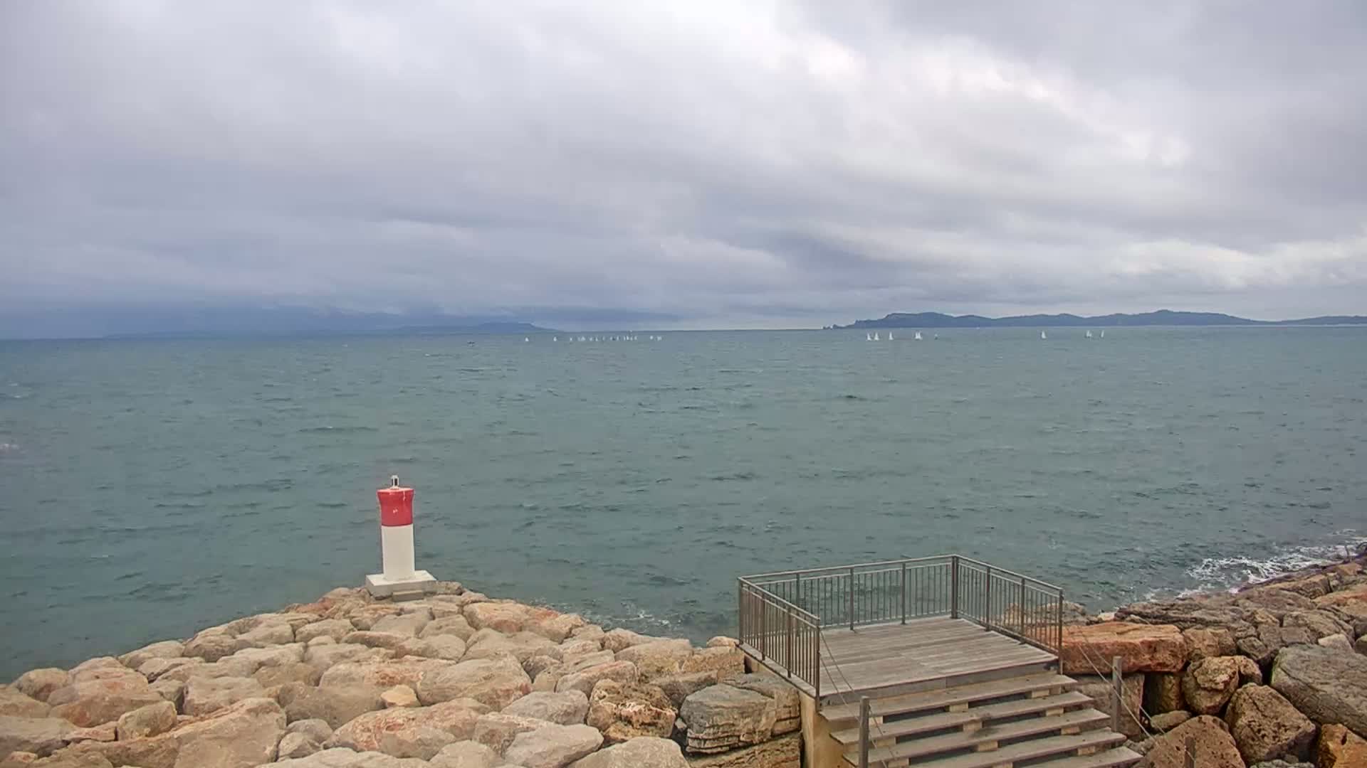 A rocky shoreline with a small wooden viewing platform and a red and white marker buoy overlooks a calm sea with distant sailboats under a cloudy sky.