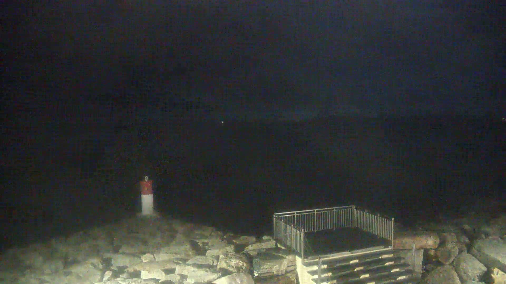A nighttime view of a rocky shoreline with a small fenced-in area and a small lighthouse, under a dark sky.