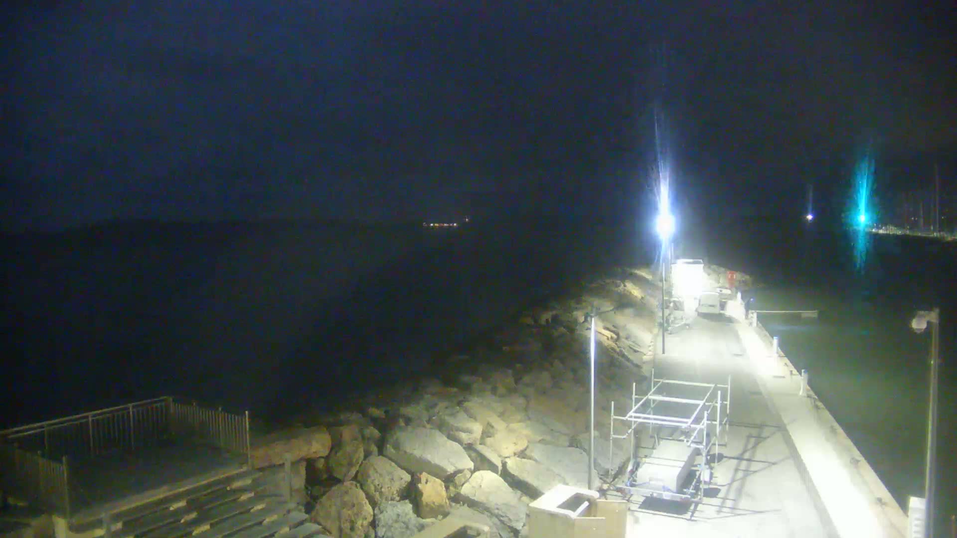 A nighttime view of a dark, calm body of water next to a stone seawall and paved walkway with some construction equipment, lit by several artificial lights.