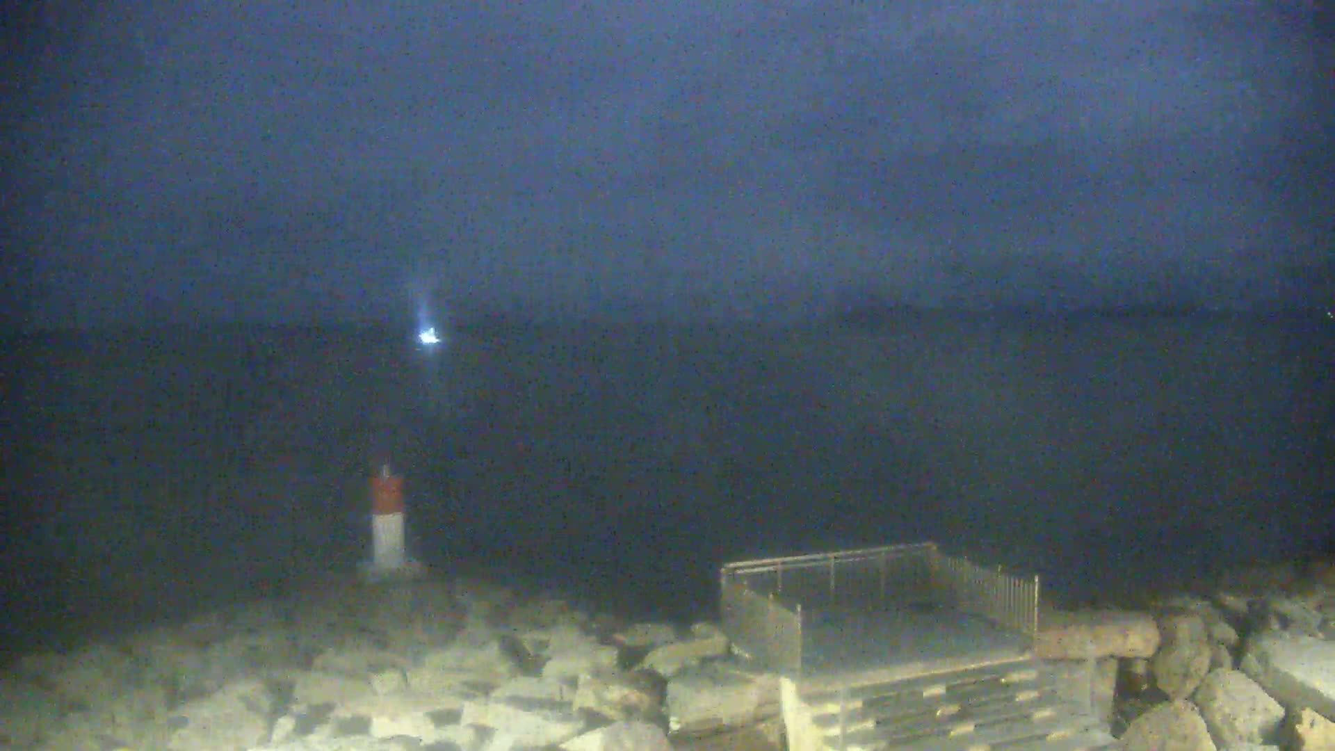 A dark, nighttime scene shows a body of water with a small light in the distance, a short, marked post near the shore, and a metal railing structure on a rocky outcrop.