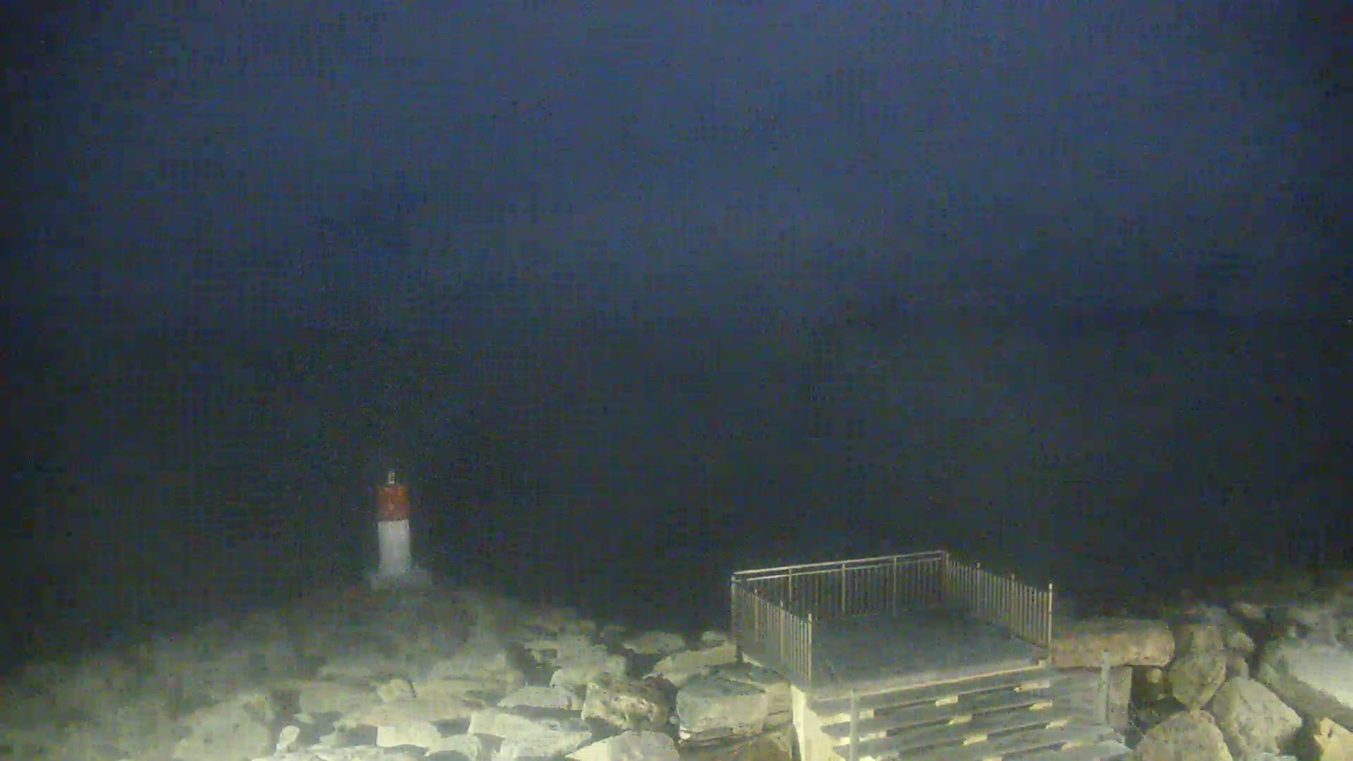 A dark, nighttime scene shows a small, white-and-red striped marker post near a wooden platform with metal railings, all situated on a rocky shoreline overlooking a dark body of water under a mostly clear, dark sky.