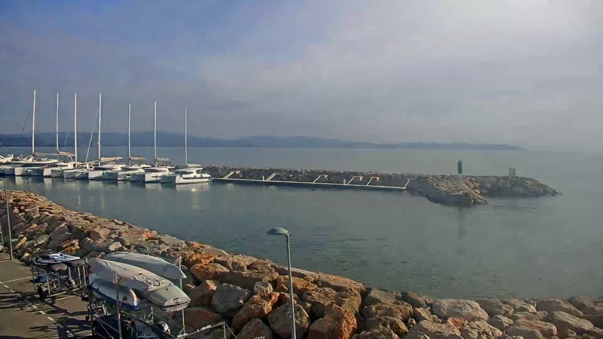 A calm body of water with numerous sailboats docked near a rocky shoreline under a hazy, sunny sky.