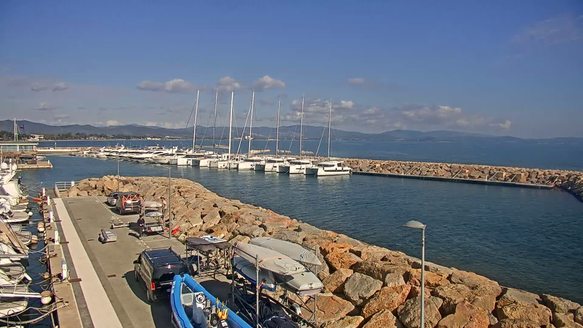 A marina filled with sailboats and motorboats is situated on a calm, sunny day, with mountains visible in the distance.