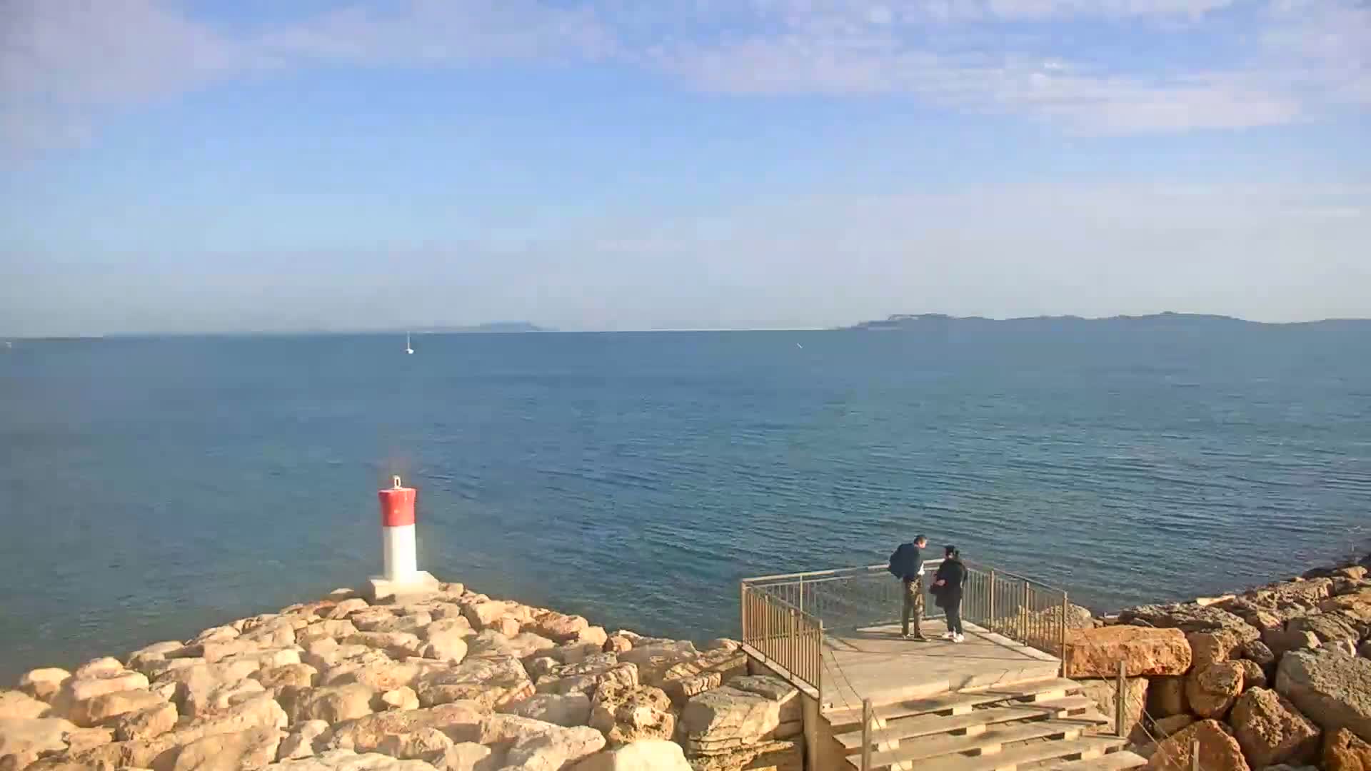 A rocky shoreline with a small lighthouse and viewing platform overlooks a calm sea under a mostly clear sky, with two people standing on the platform.