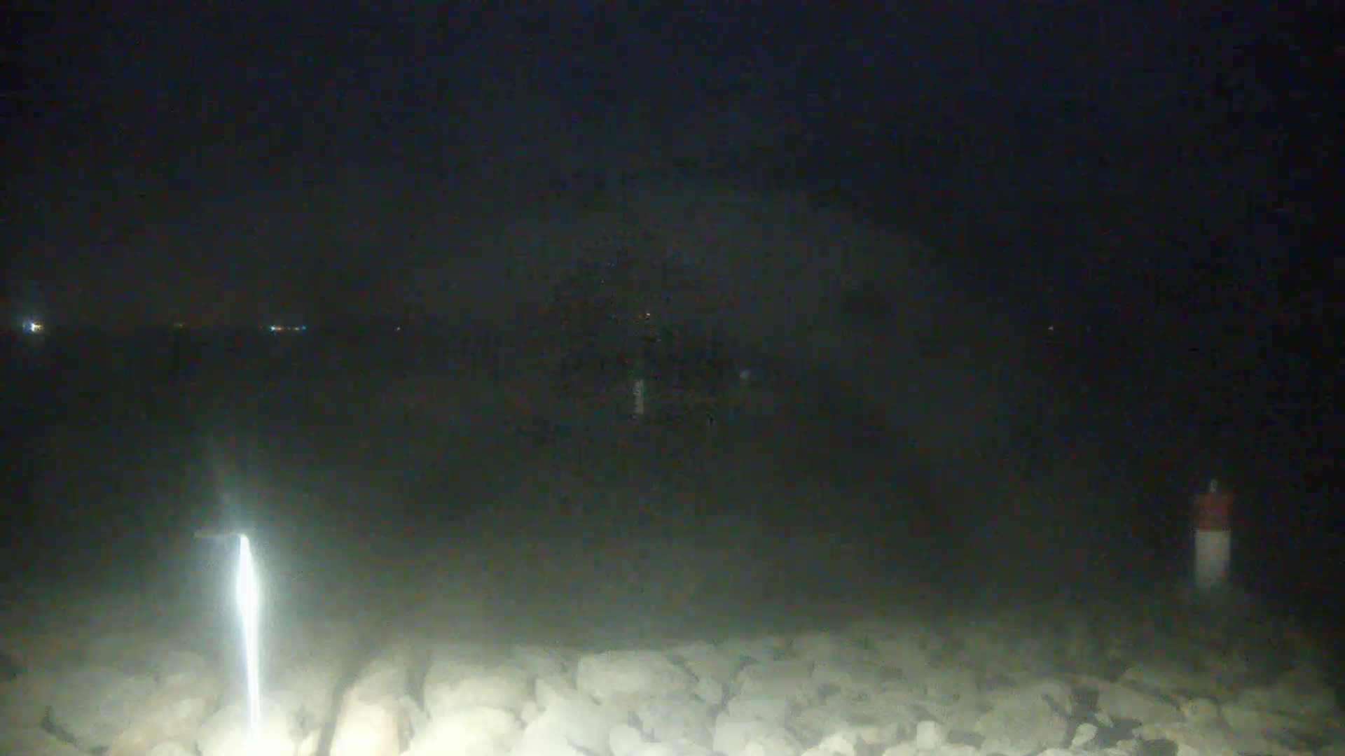 A bright light illuminates a rocky shoreline at night, with a dark, possibly foggy, background showing distant lights.