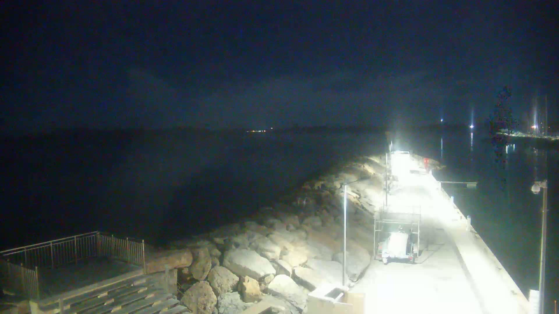 A nighttime view of a rocky shoreline and a lit pier extending into a calm, dark body of water under a clear, dark sky.