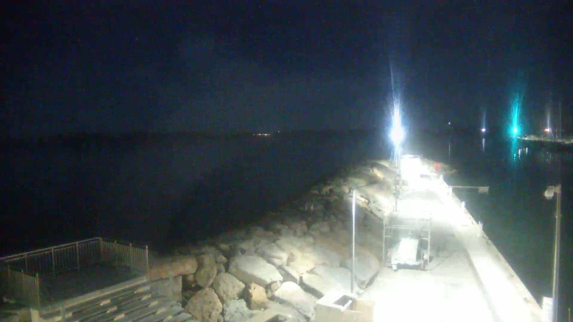 A nighttime view of a rocky breakwater and illuminated dock next to a calm body of water under a clear, dark sky.