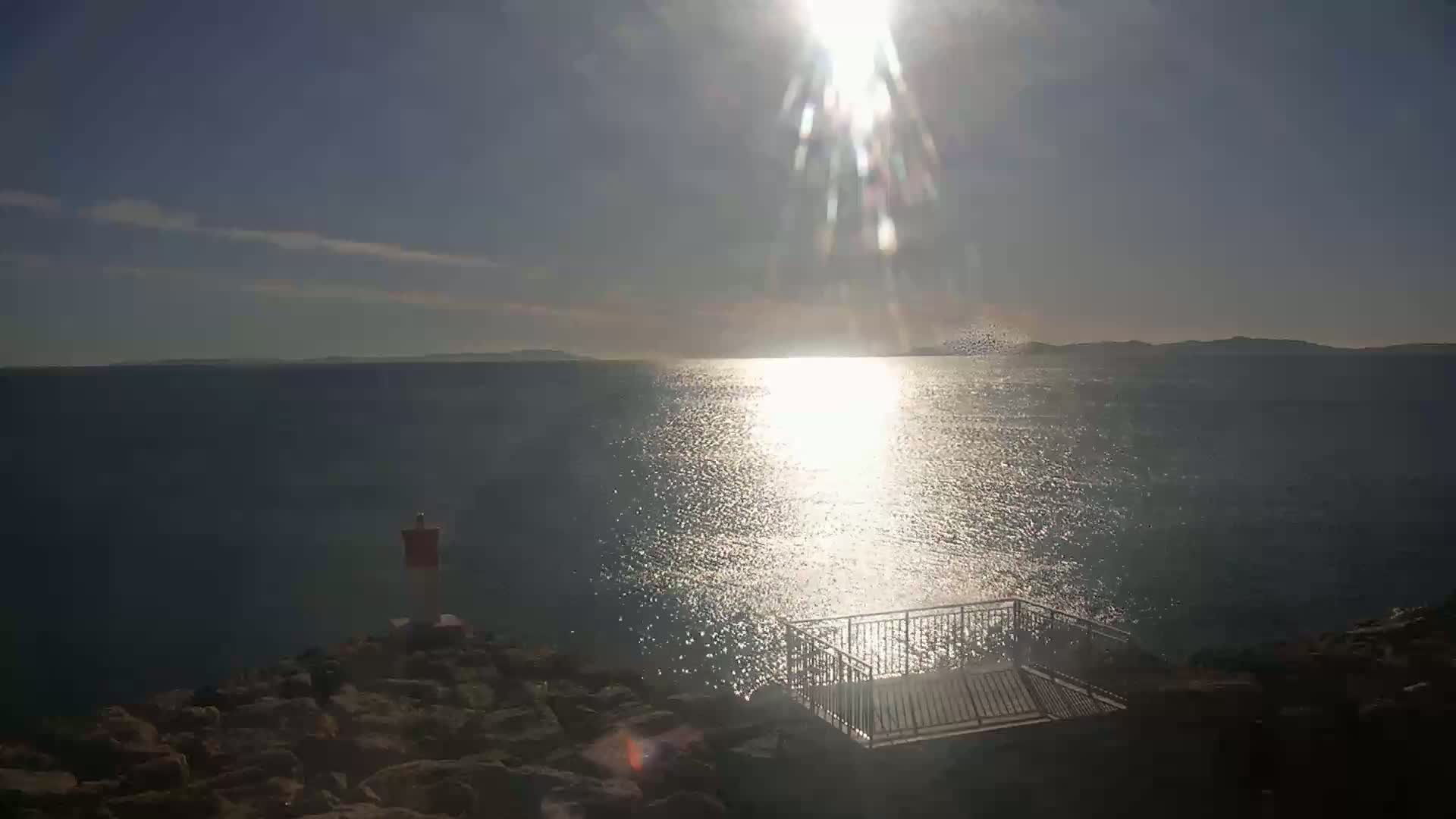 A bright, sunny day reveals a calm body of water reflecting the sun's glare, with a small metal railing and stairs leading down to the rocky shoreline.