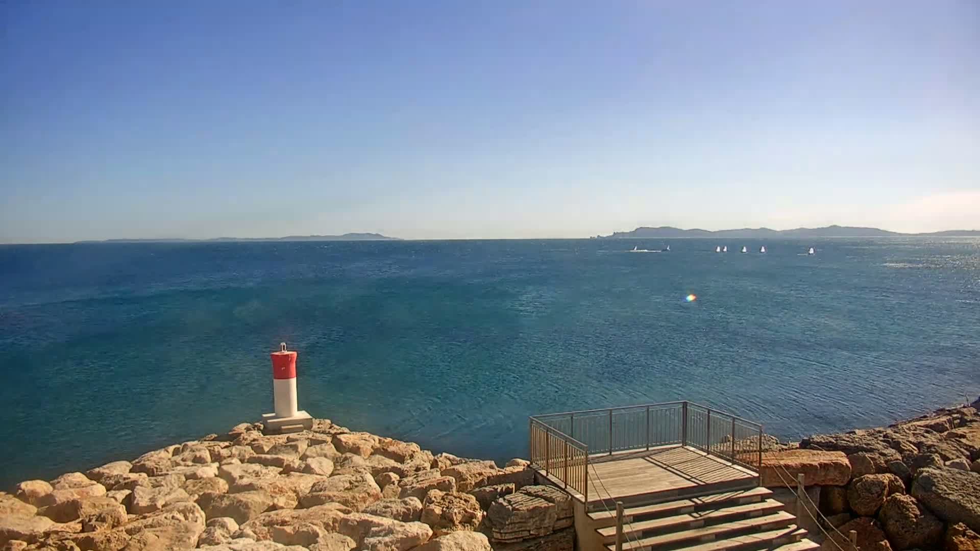 A rocky shoreline with a small wooden viewing platform overlooks a calm, blue sea where several small boats sail under a clear sky.