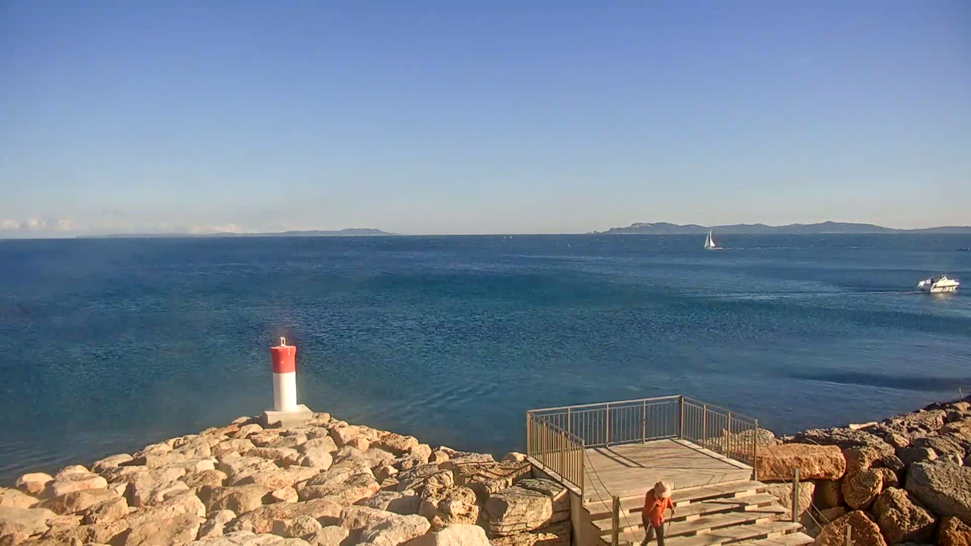 A person stands on wooden steps overlooking a calm blue sea, a small red and white lighthouse visible to the left, and distant islands under a clear blue sky.