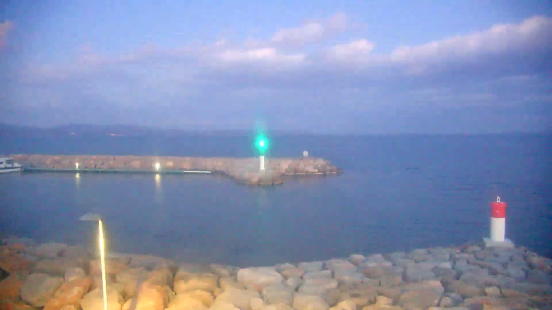 A small harbor with a lighthouse and a breakwater is visible under a mostly cloudy sky.