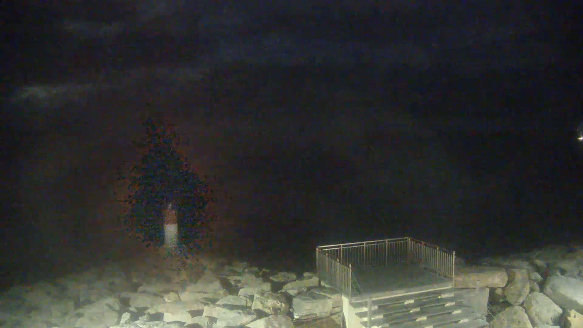 A nighttime view of a rocky shoreline with a small metal railing and stairs leading down to the water under a dark, cloudy sky.