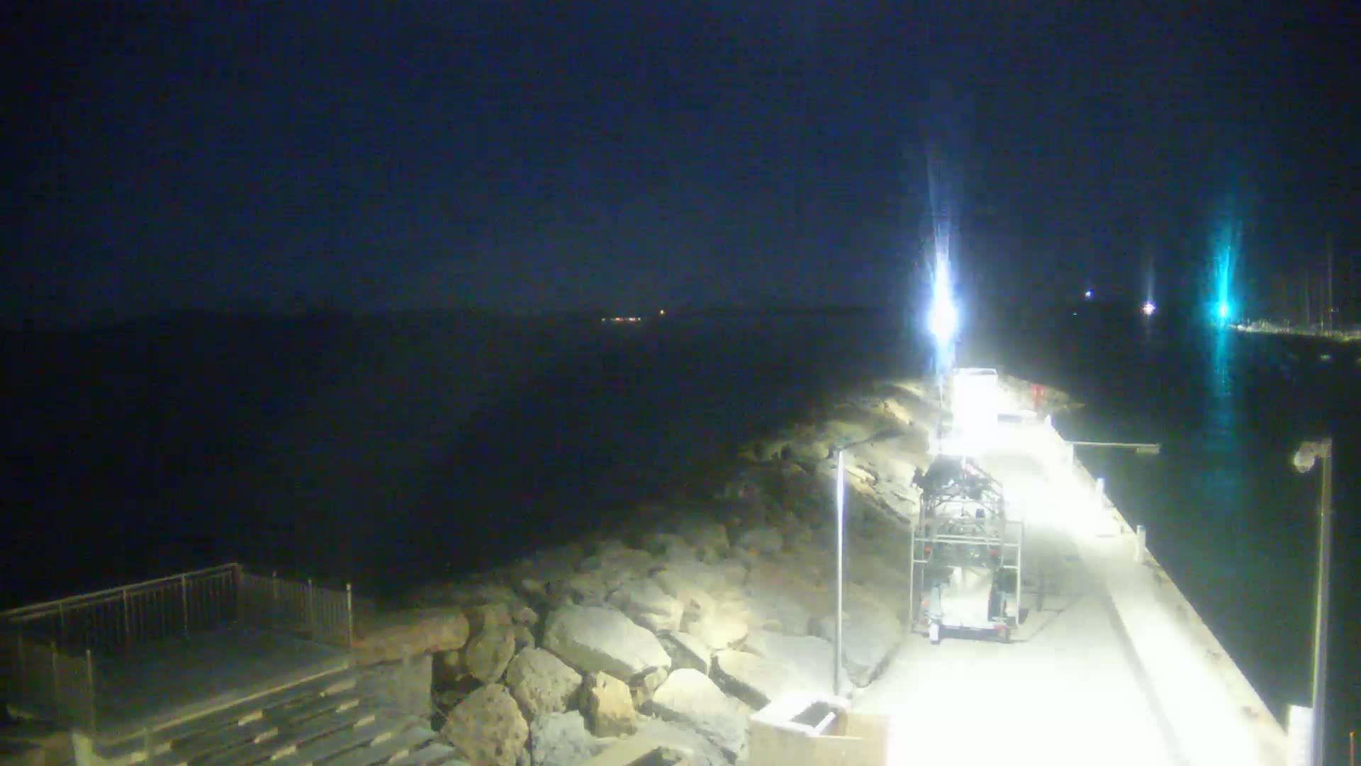 A nighttime view of a rocky shoreline and a paved walkway, illuminated by lights, with a trailer and other vehicles parked along the walkway and calm dark water visible in the background.