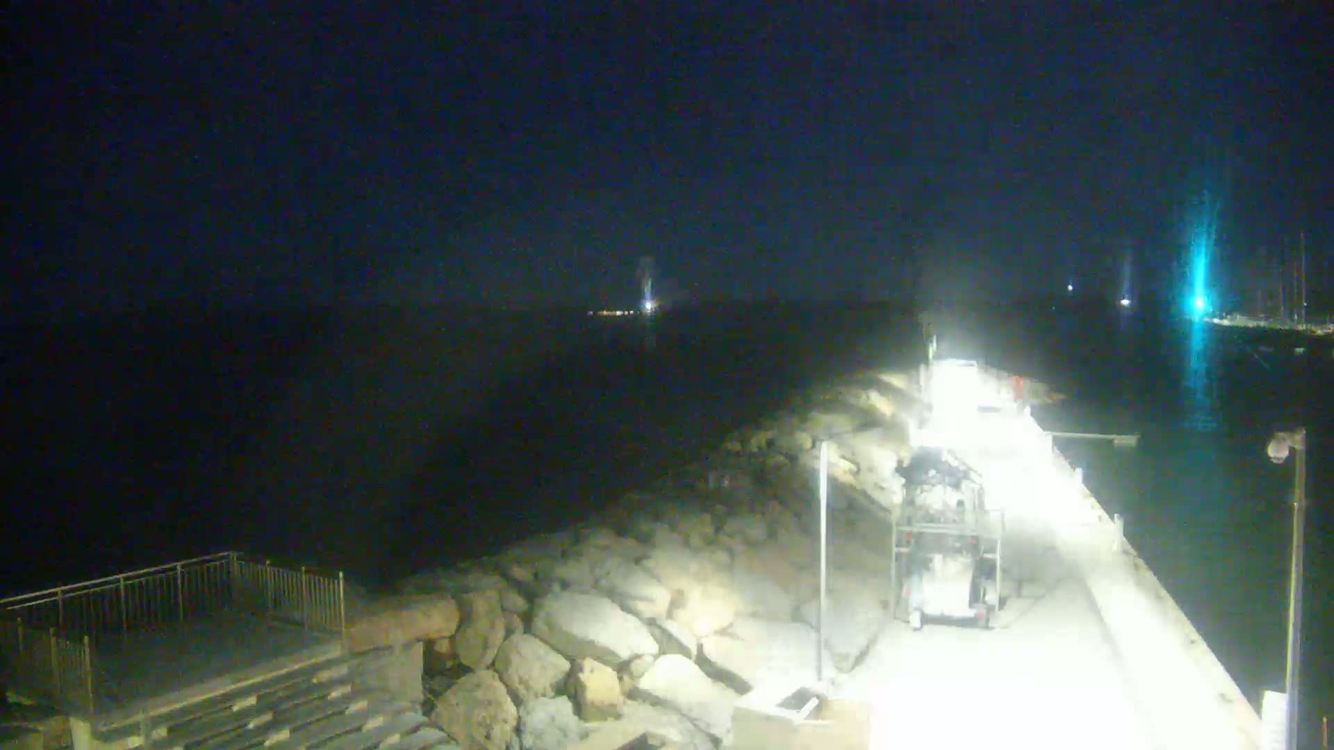 A nighttime view of a rocky shoreline and a paved walkway with vehicles, illuminated by lights, overlooking dark water.