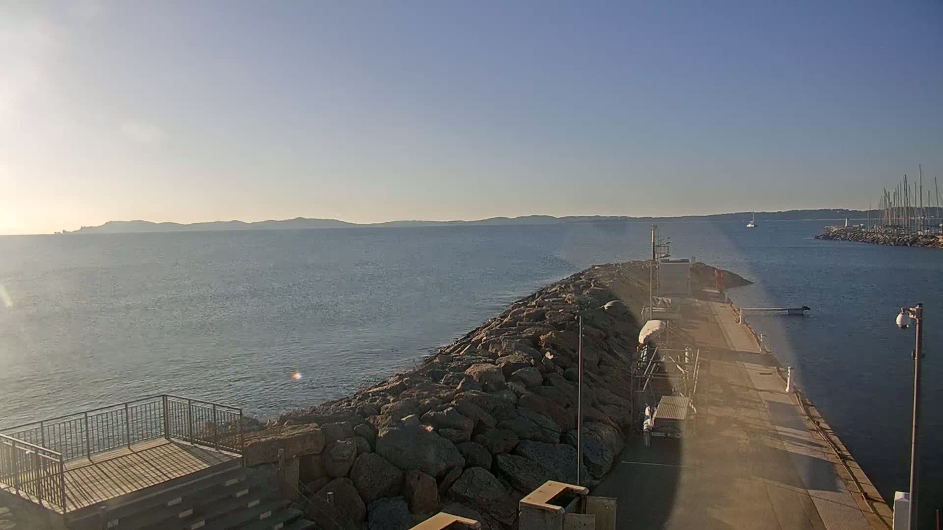 A rocky breakwater extends into a calm, blue sea under a clear, bright sky, with distant land visible on the horizon.