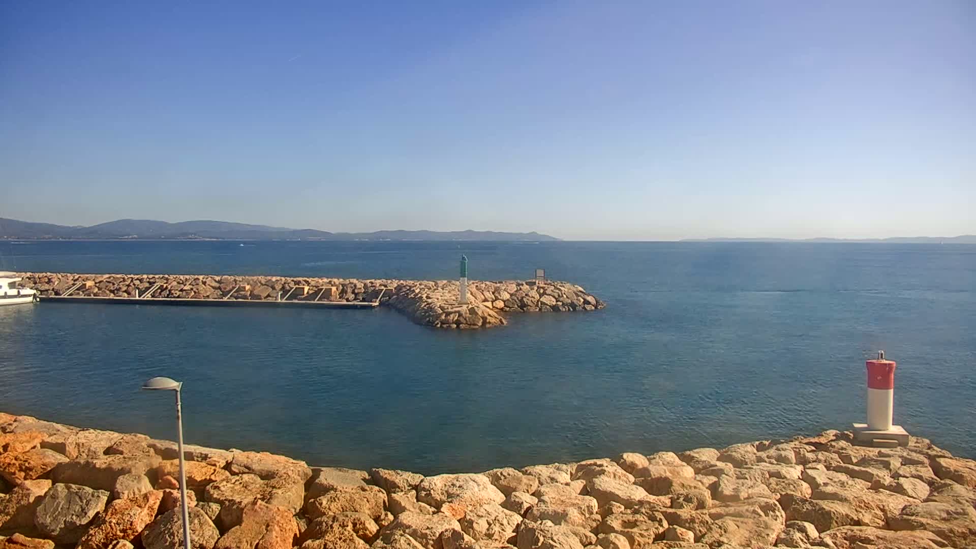 A calm, blue sea stretches to a distant mountain range under a clear blue sky, with a small harbor protected by a rock jetty featuring a small lighthouse.
