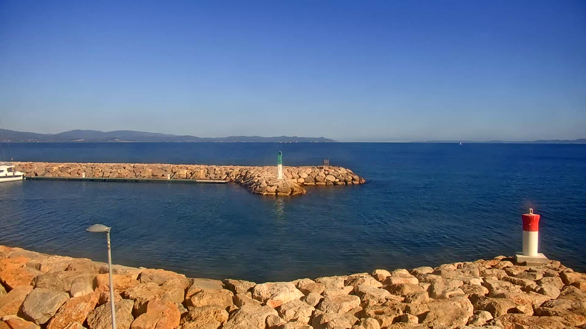 A calm, clear day reveals a rocky harbor with a small boat docked, a lighthouse, and distant mountains across a blue sea.