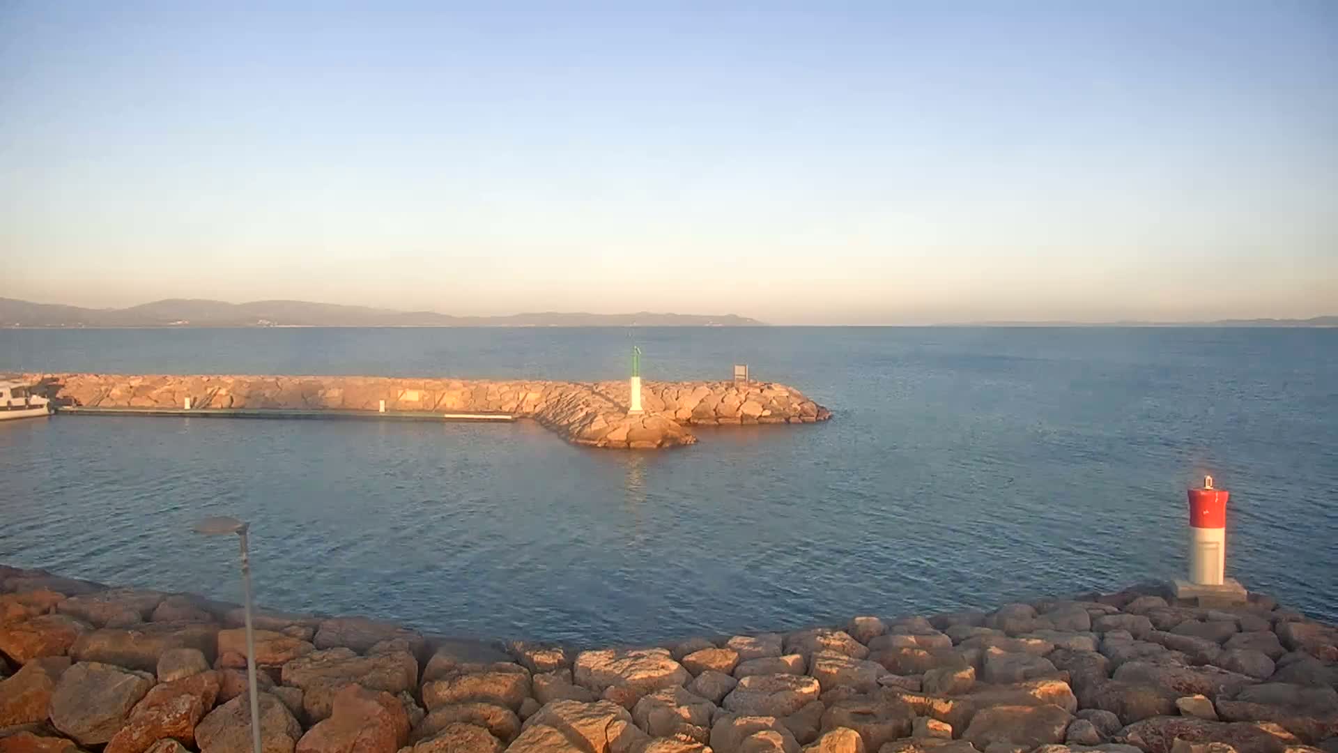 A calm, sunlit body of water is partially enclosed by a rocky jetty, with a small lighthouse and a distant mountain range visible on a clear day.