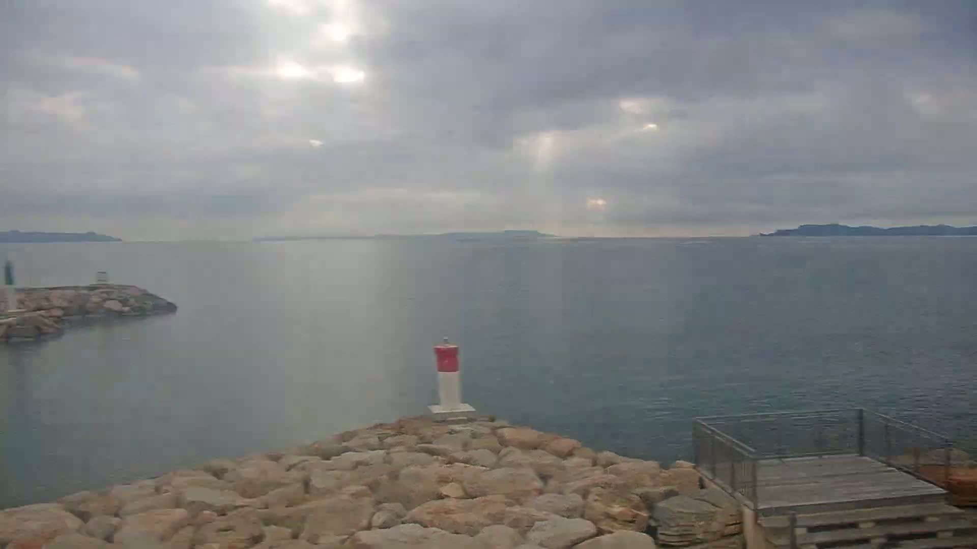 A rocky coast with a small lighthouse overlooks a calm sea under a cloudy sky with sunbeams breaking through.