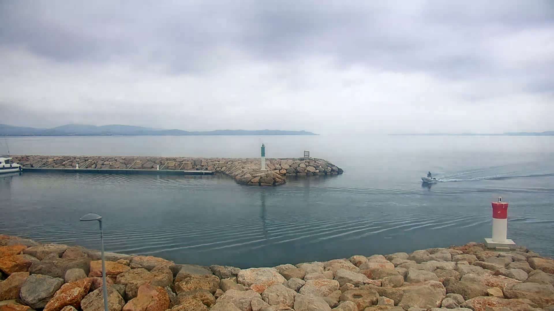A small motorboat travels across calm, gray water past a rocky jetty with a lighthouse under an overcast sky.