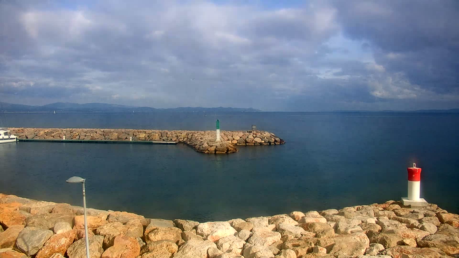 A partly cloudy day reveals a tranquil harbor scene with a small boat docked near a rock jetty, a lighthouse marking a corner, and a distant mountain range visible across the calm water.