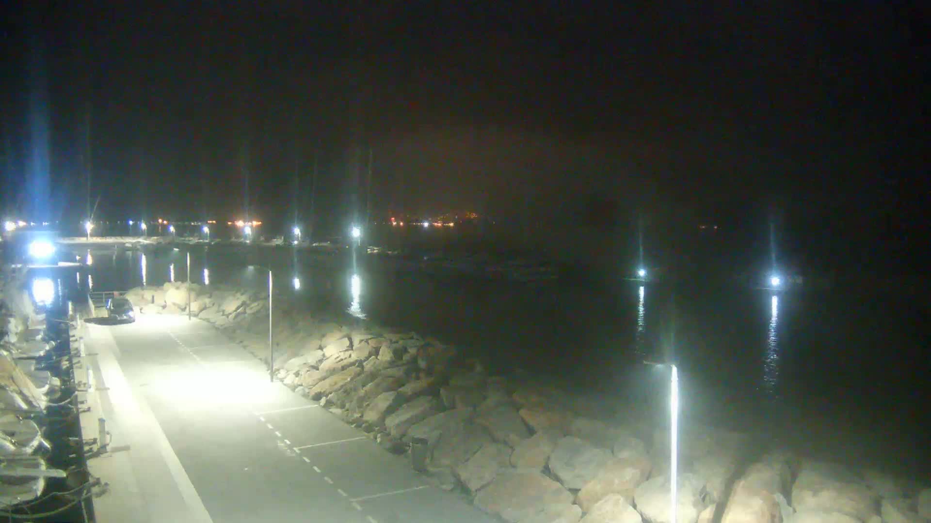 A nighttime view of a paved waterfront area with lights lining a rocky embankment, overlooking a calm body of water with boats moored in the distance.