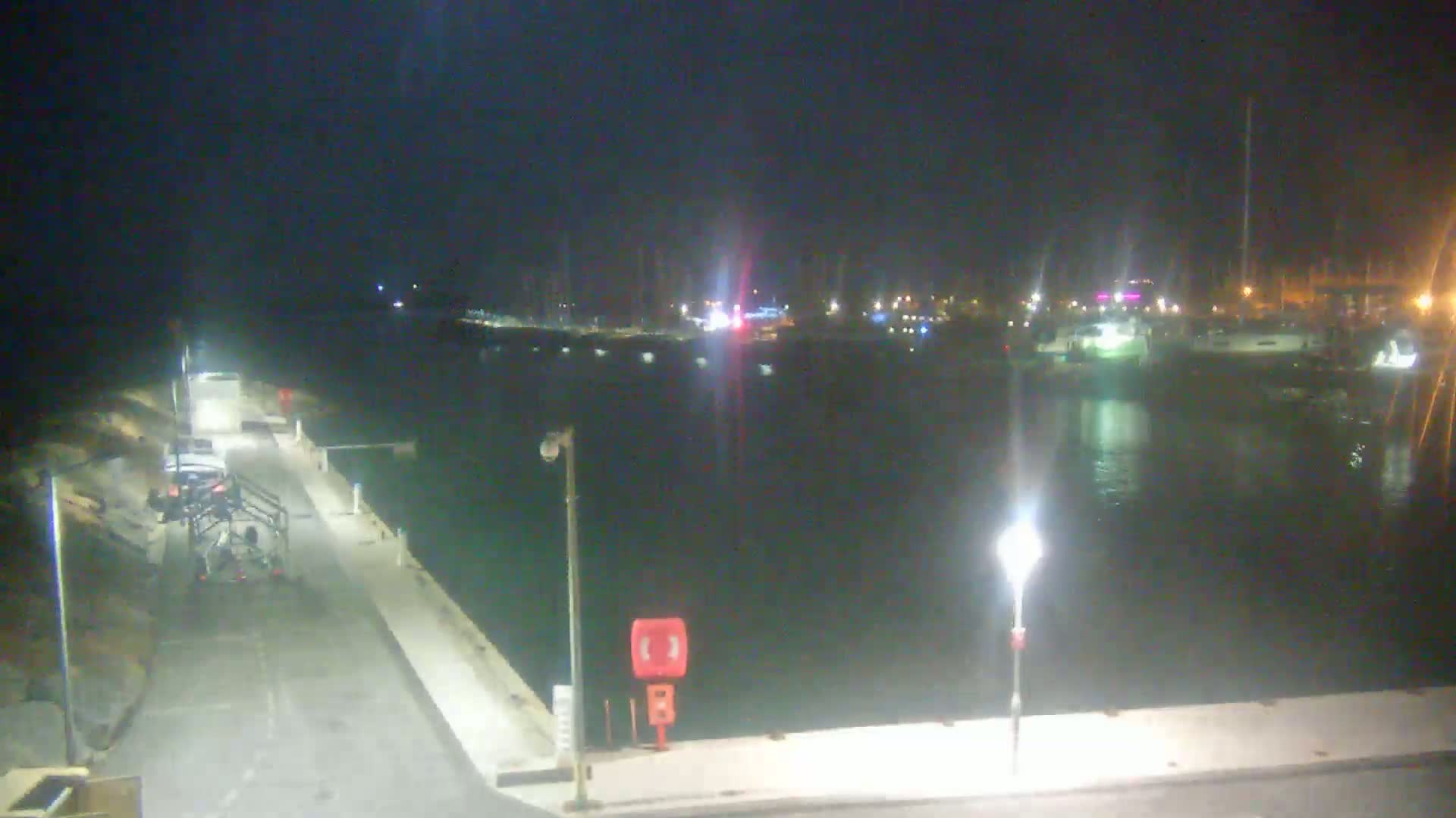A nighttime view of a harbor with docked boats and a paved walkway leading to a parking area, illuminated by streetlights.
