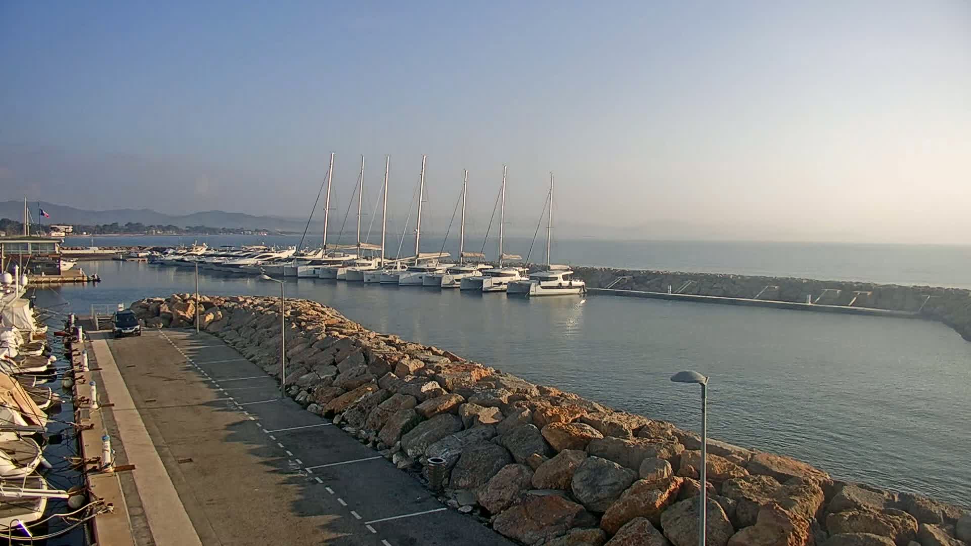 A calm, hazy harbor scene shows a row of sailboats moored near a rocky breakwater, with a parking lot and additional boats visible in the foreground.