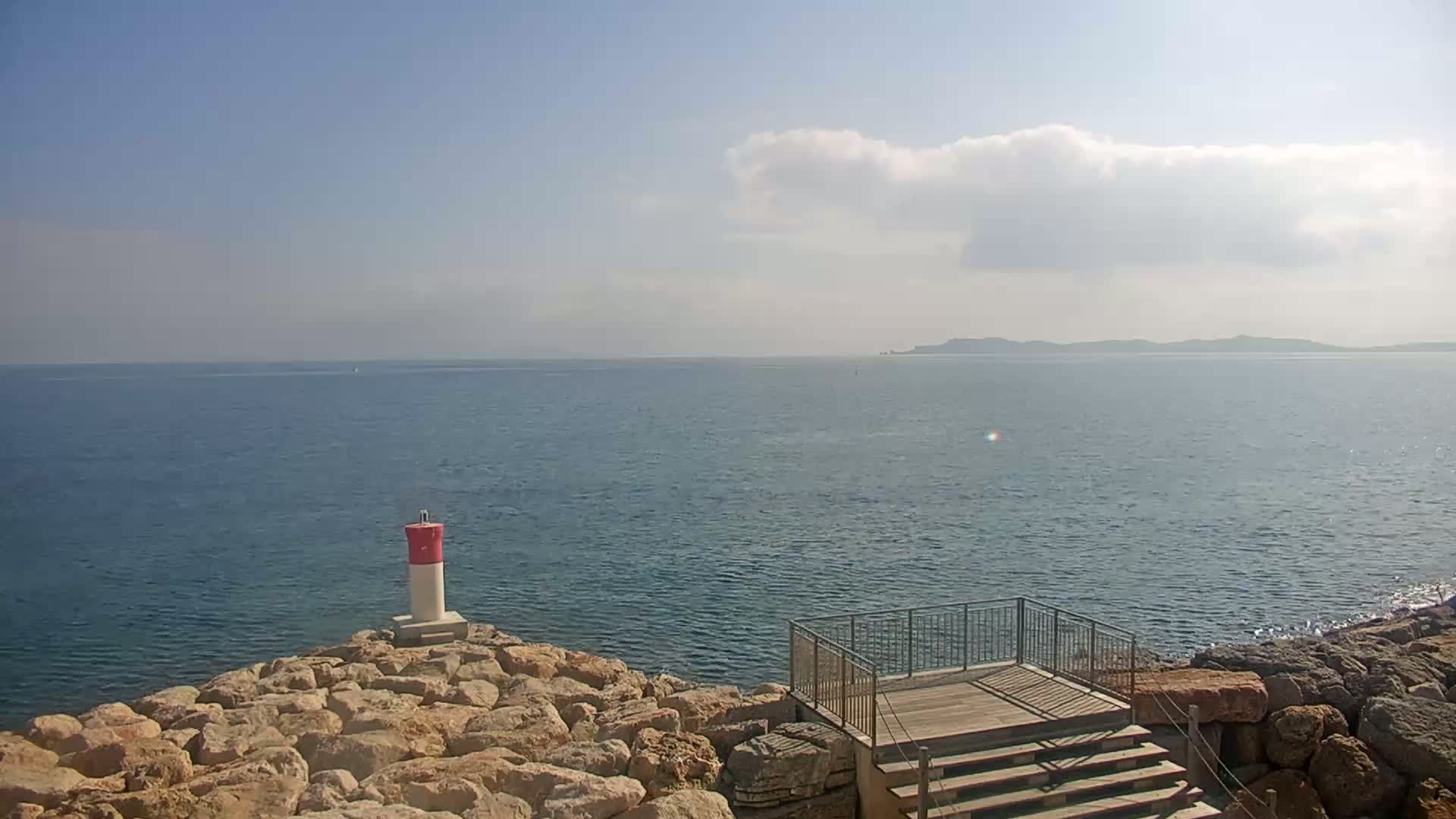 A small, red and white lighthouse stands on a rocky outcrop overlooking a calm sea under a partly cloudy sky.