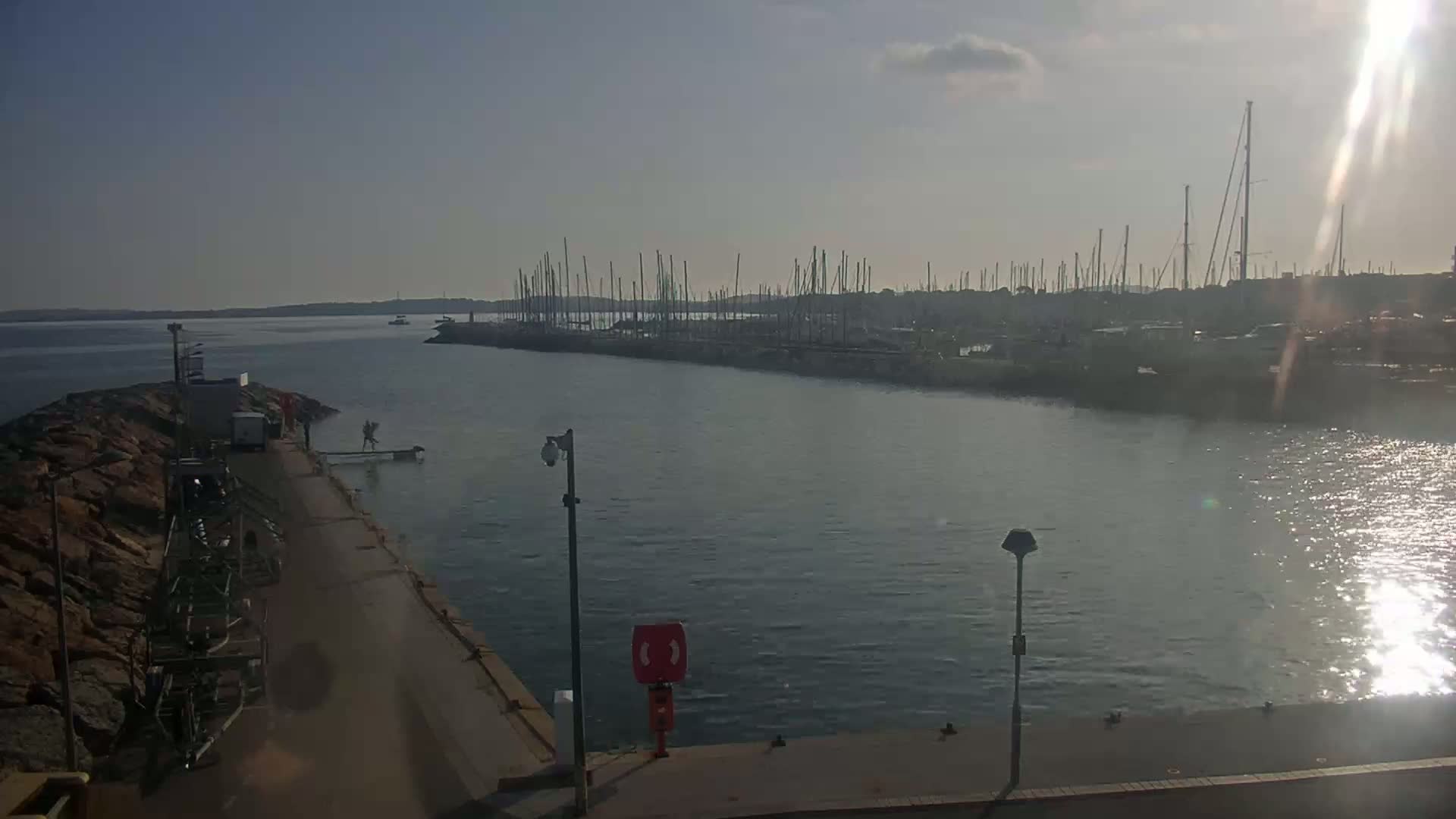 A calm, sunny day at a marina shows numerous sailboats docked, viewed from a quayside with a rocky outcrop.