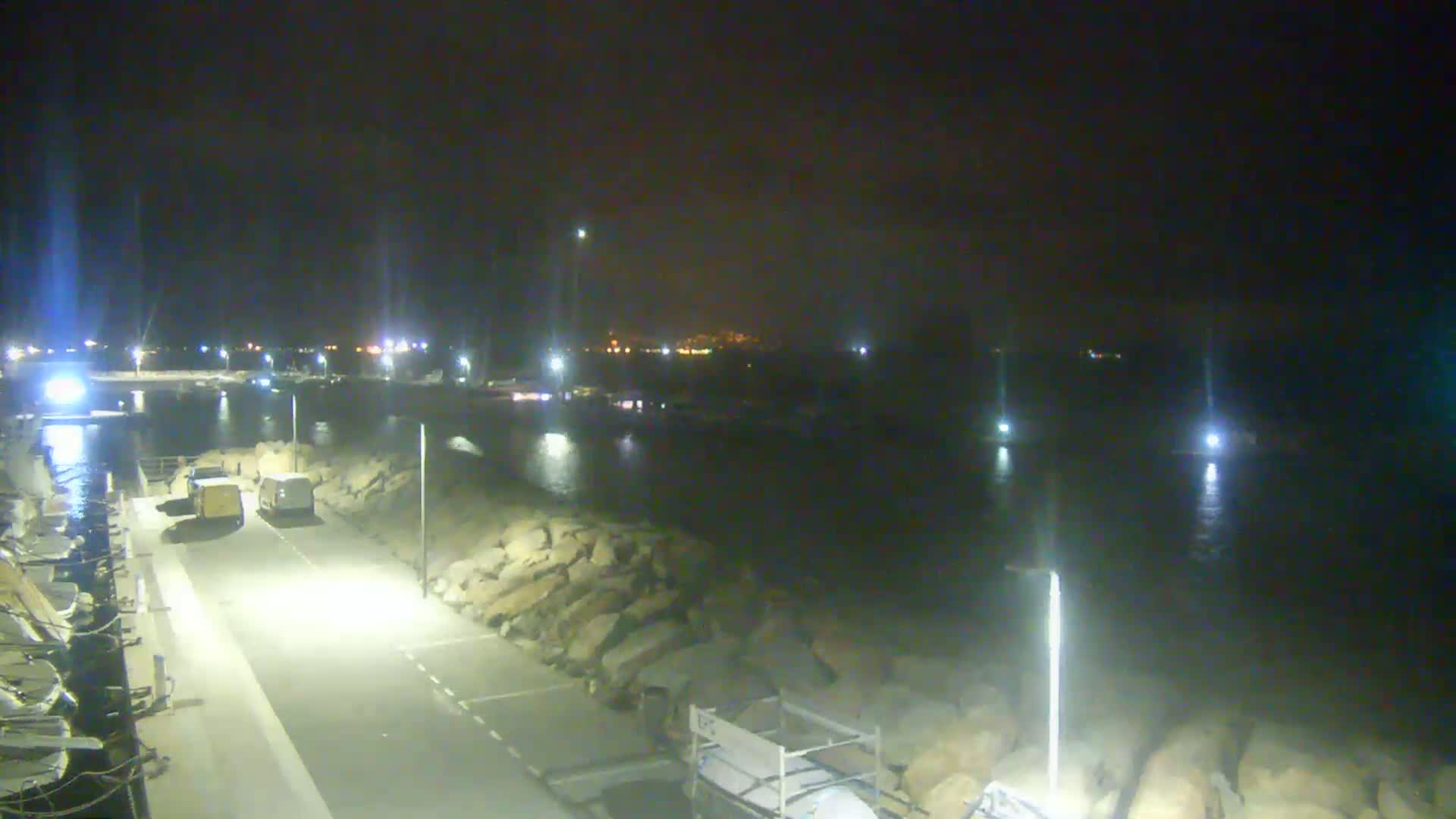 A nighttime view of a harbor with boats, a roadway, and rock barriers, under clear skies.