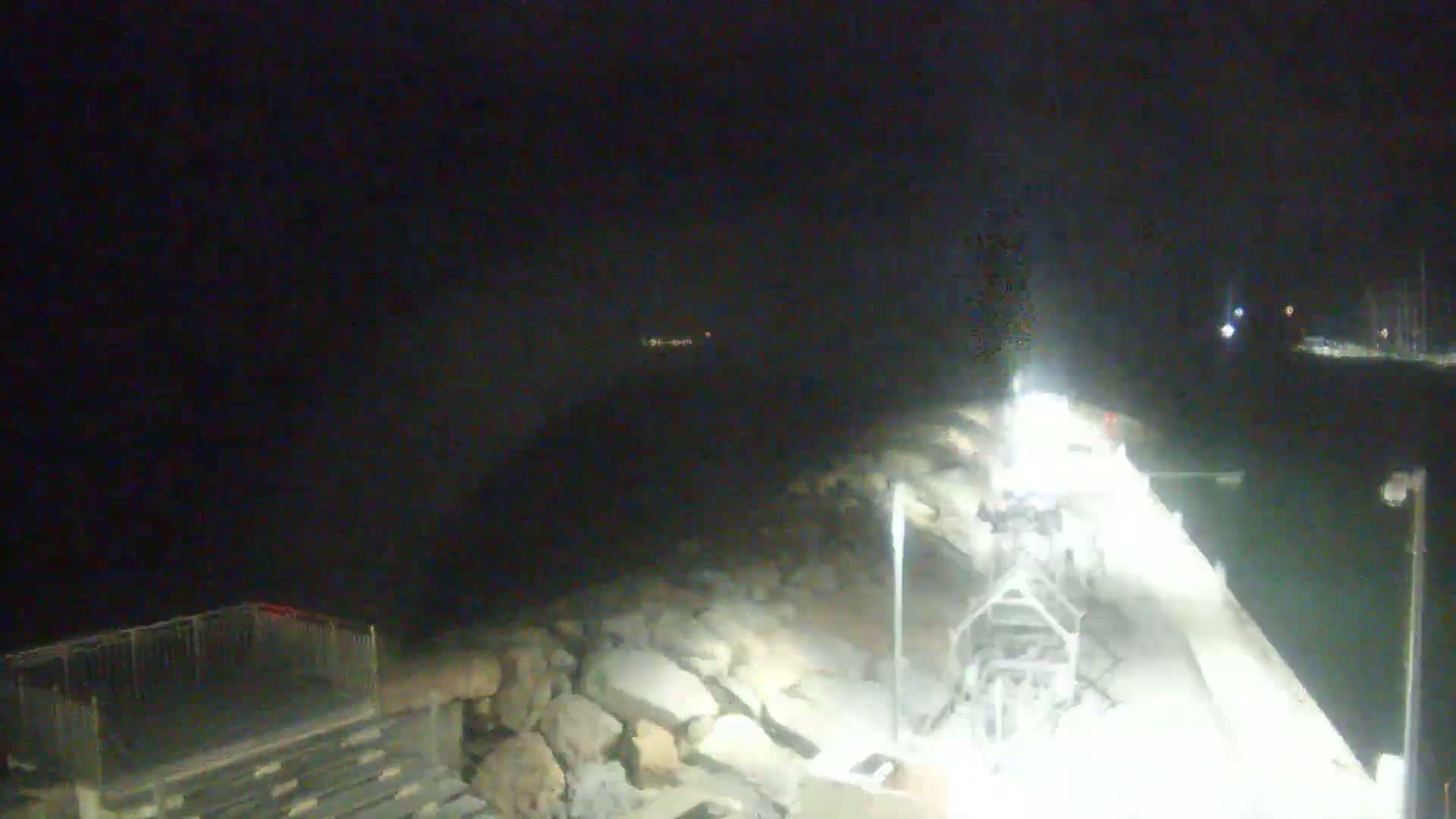 A brightly lit pier extends over dark water at night, with a rocky shoreline and distant lights visible.