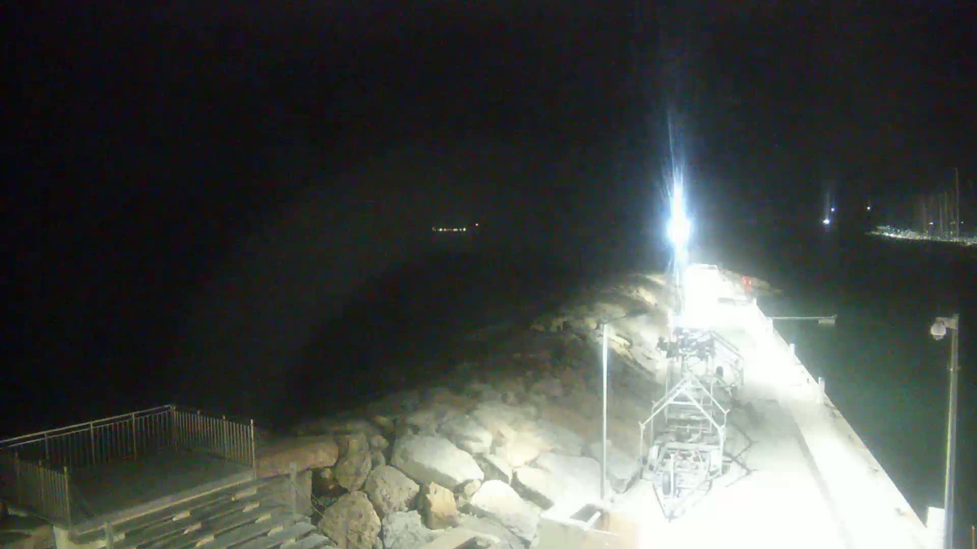 A brightly lit pier extends into dark water at night, with a rocky shoreline and distant lights visible.