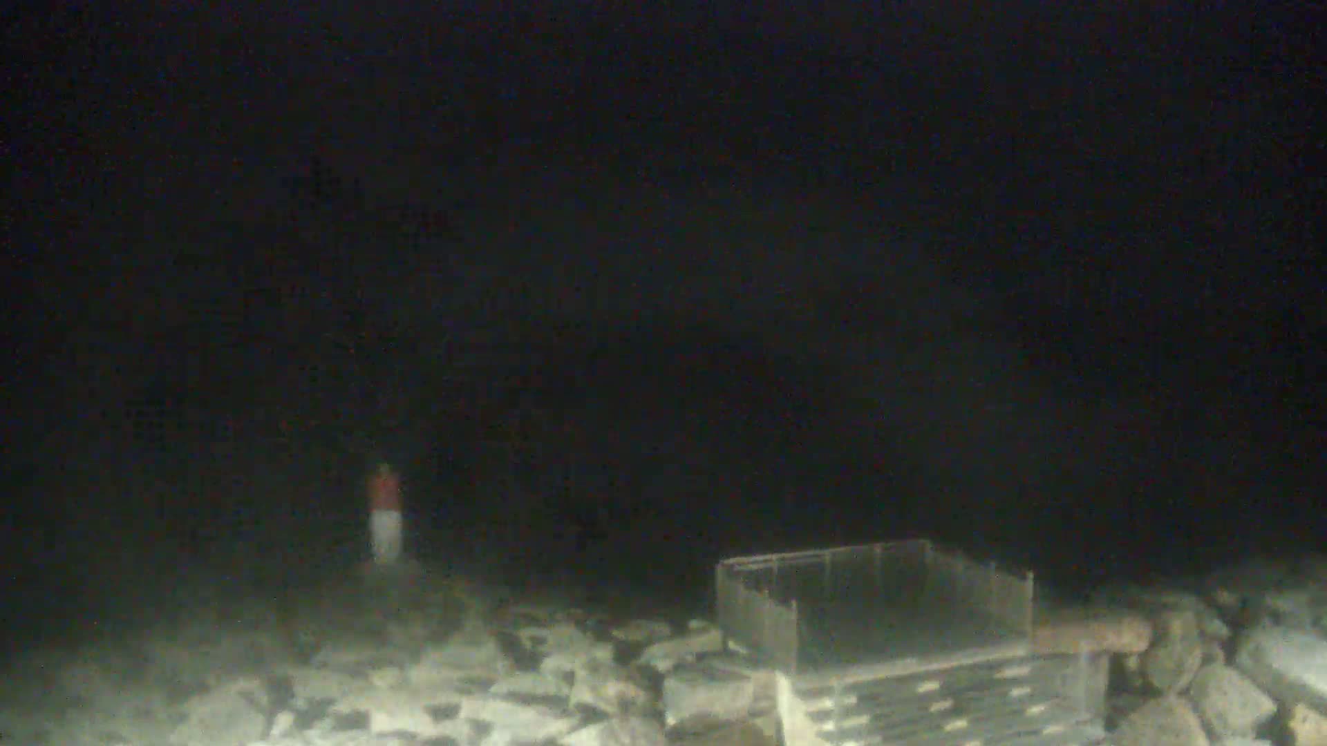 A person stands near a metal structure on a rocky shore at night under a dark, overcast sky.