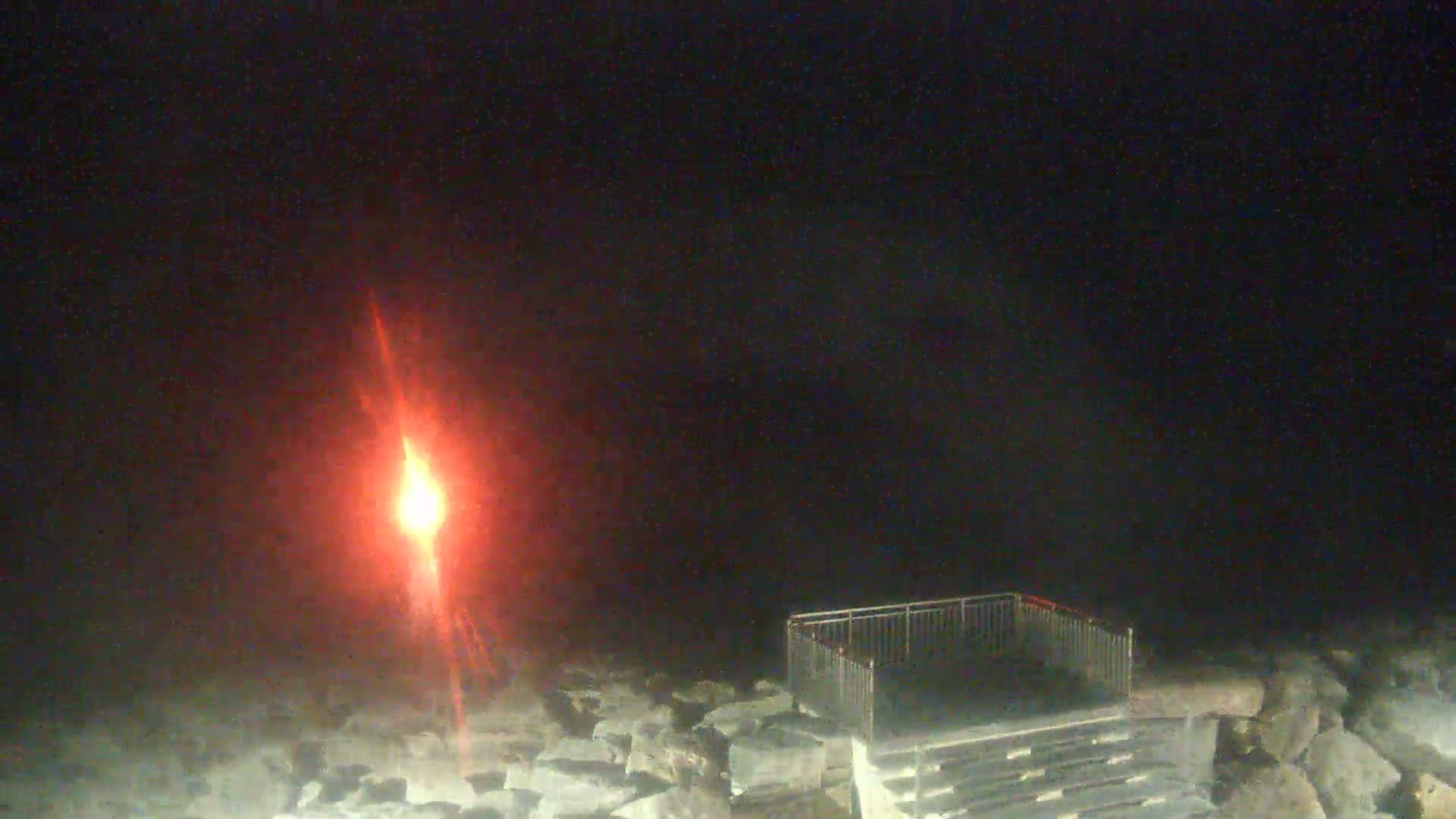 A bright red light shines in the dark night sky over a rocky shoreline with a metal railing and steps.