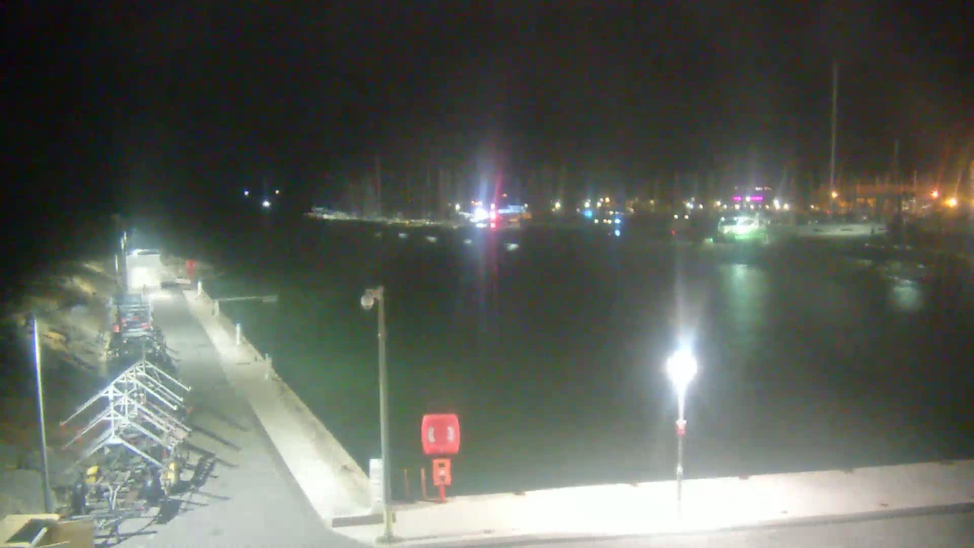 A nighttime view of a harbor with boats docked, lit by several lights, and a walkway with equipment along the side.
