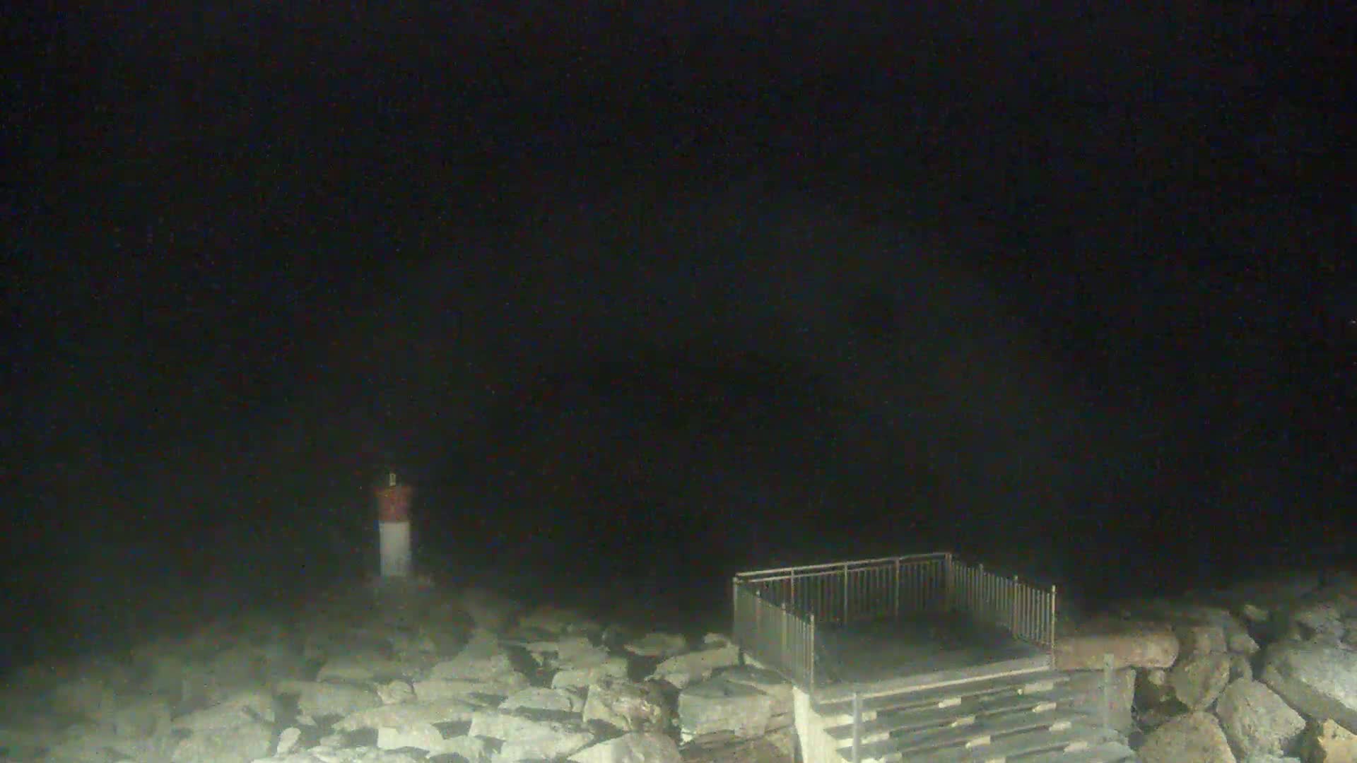 A small, nighttime seaside scene shows a short set of stairs leading to a gated platform beside a small lighthouse on a rocky shore under a dark, cloudy sky.