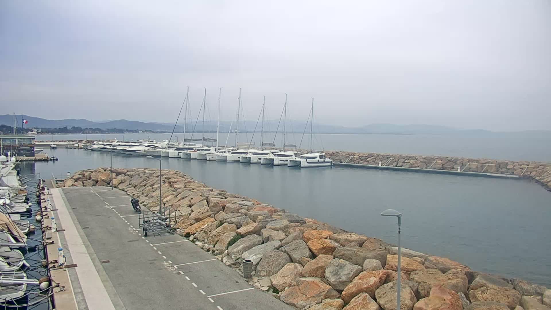 A marina with numerous yachts and sailboats is nestled within a stone breakwater, overlooking calm water under an overcast sky.