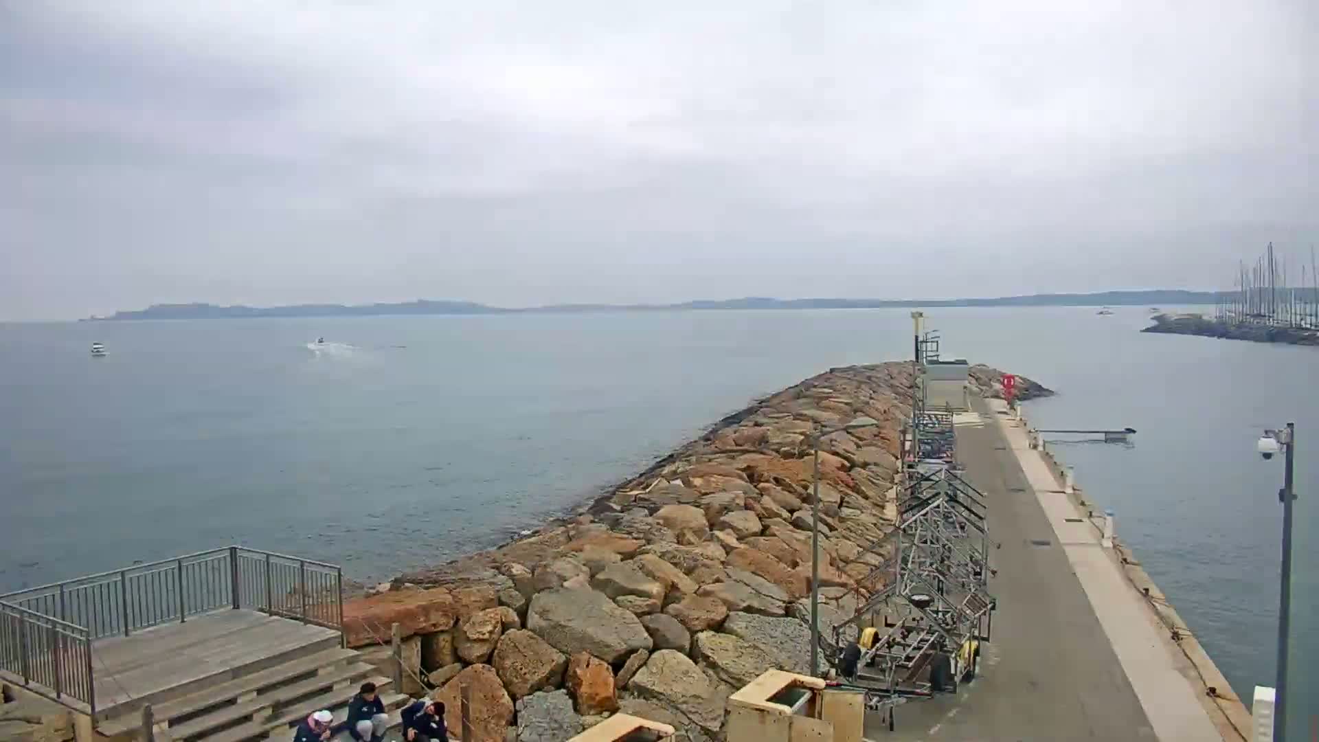 A rocky breakwater extends into a calm, gray sea under an overcast sky, with several small boats visible in the distance and a few people gathered near a viewing platform.