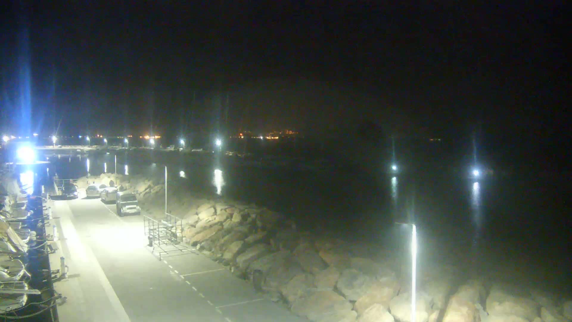 A nighttime view of a paved area next to a rocky shoreline, illuminated by several bright lights, with a few vehicles parked nearby and distant lights visible across the water.