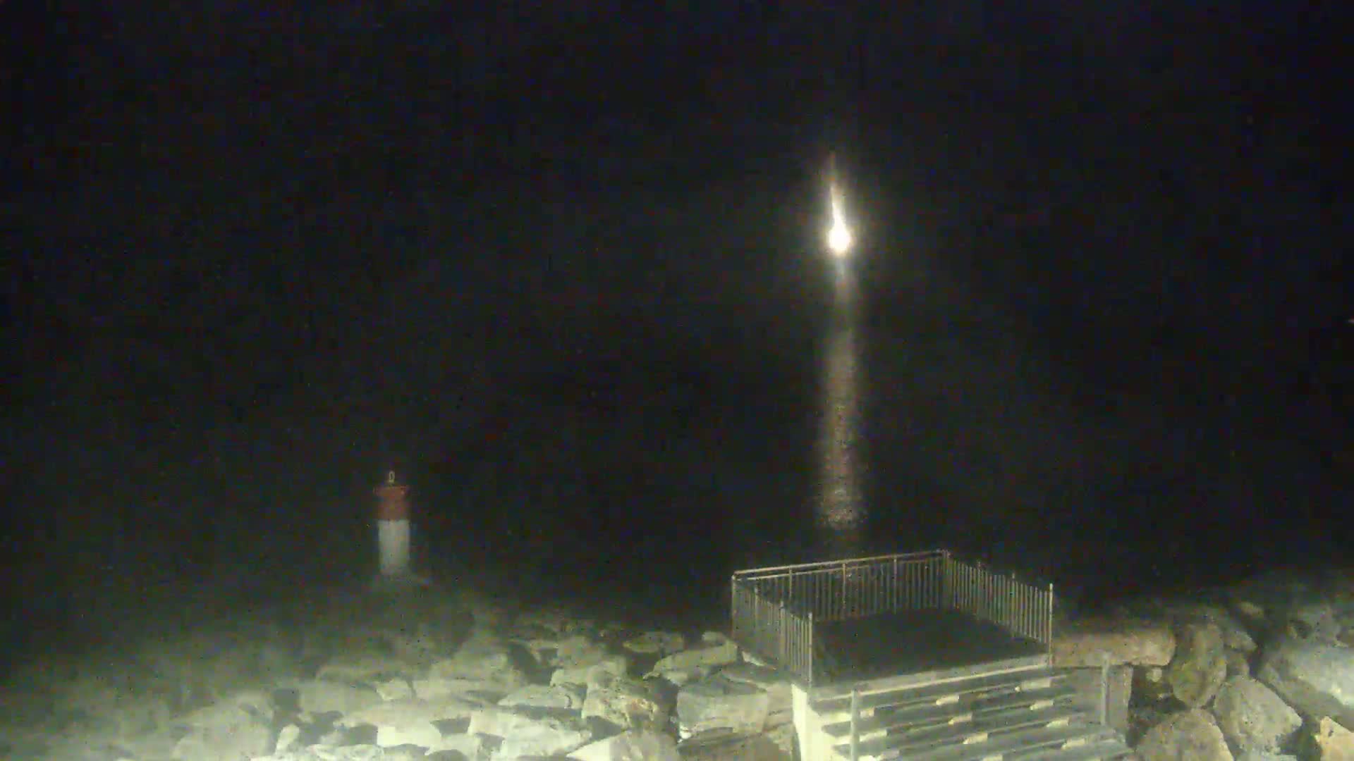 A bright light shines over dark water from a distant source, illuminating a rocky shoreline with a small structure and a marker.