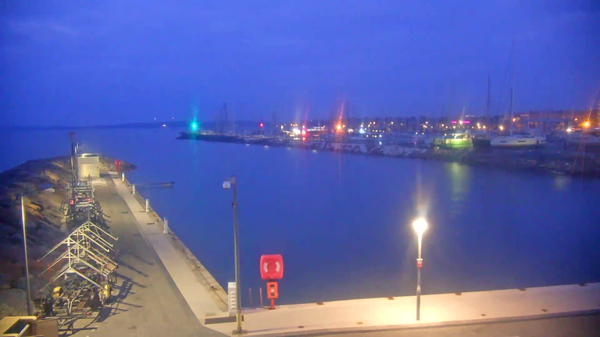 A harbor at night with calm water, under a twilight sky, showing boats moored at the far end of a pier with lights illuminating the scene.
