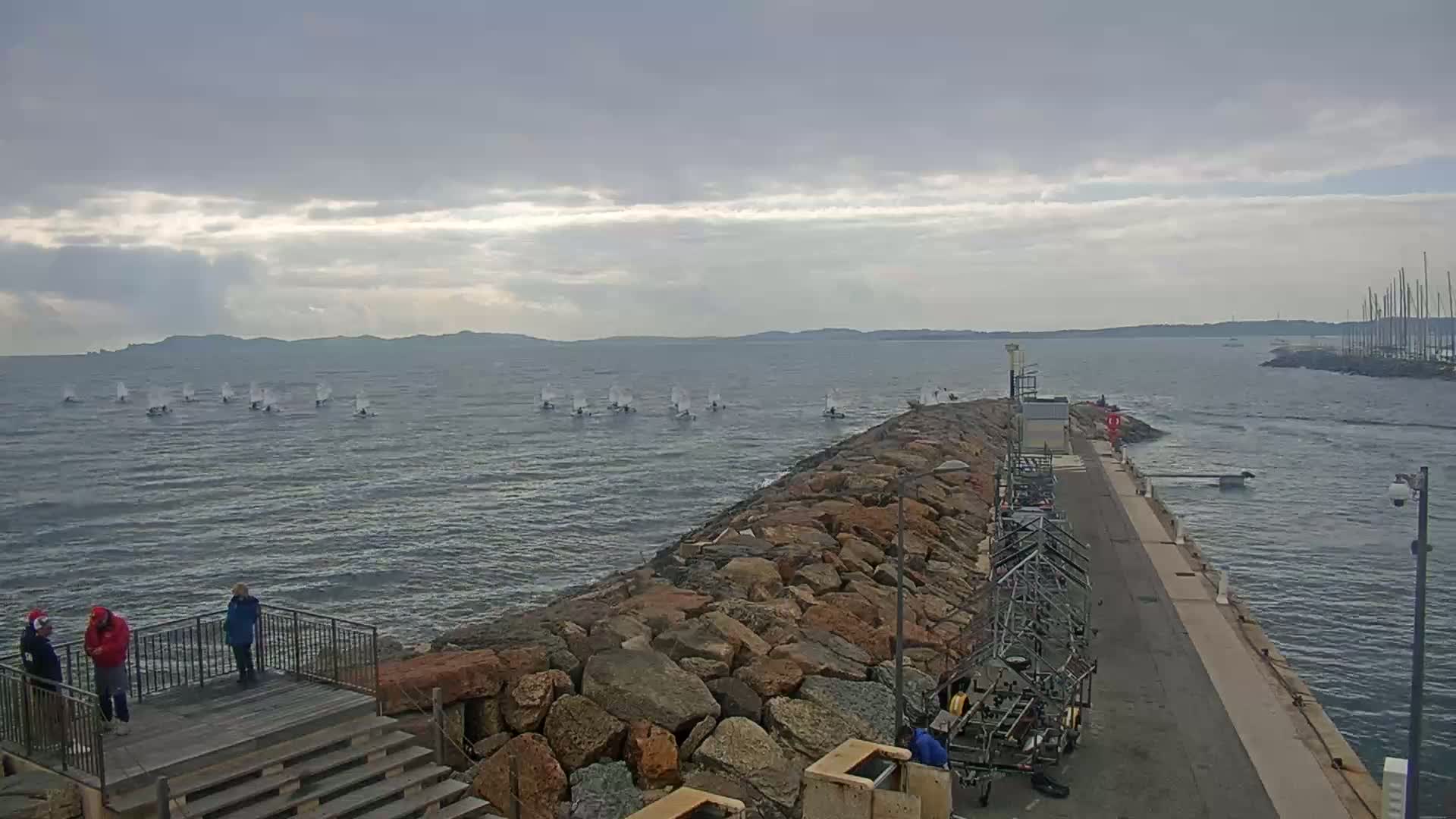 Several small sailboats are sailing on a choppy sea under a cloudy sky, alongside a rocky breakwater with people standing on a nearby viewing platform.