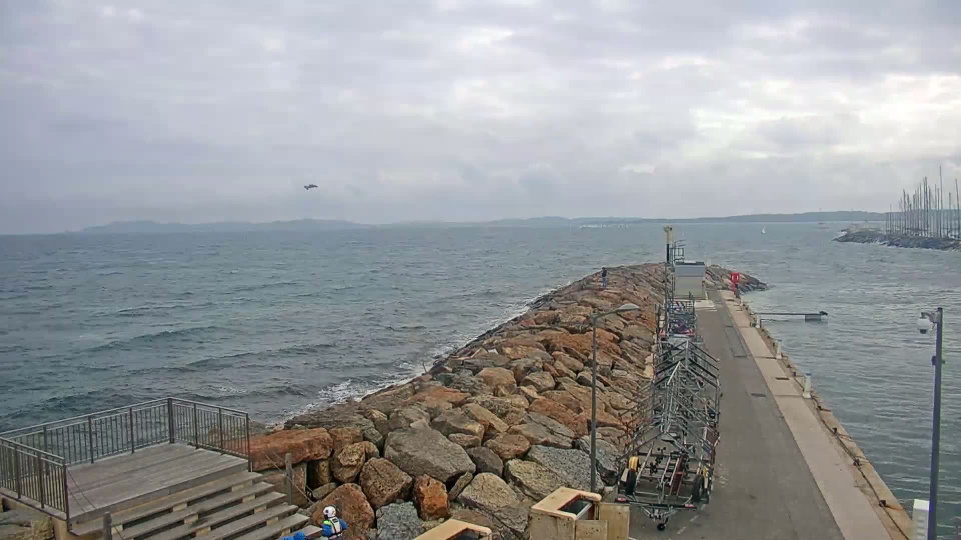 A rocky breakwater extends into a choppy sea under a cloudy sky, with a distant shoreline visible.