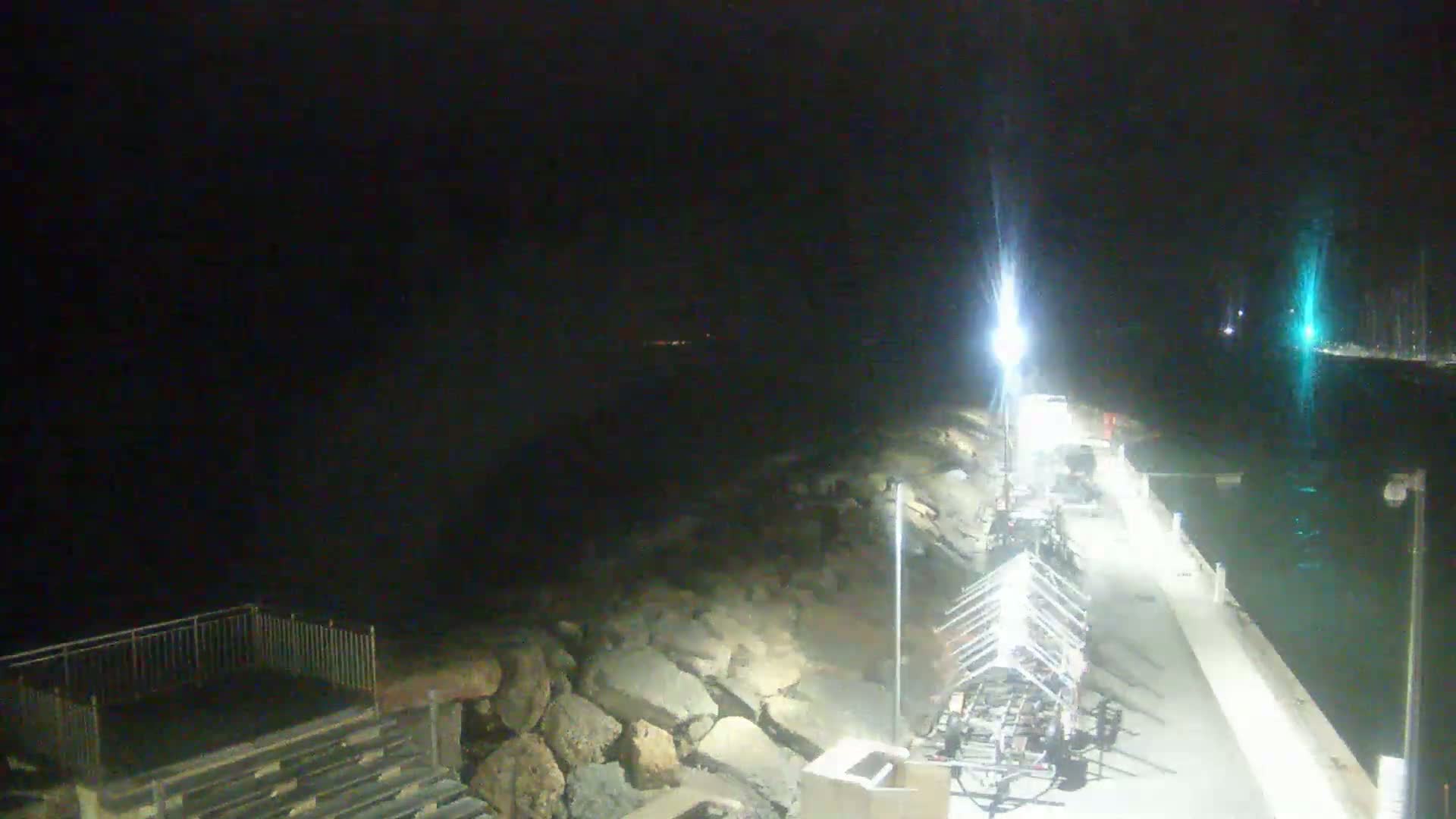 A nighttime view of a harbor with a paved walkway, rocky shoreline, and several boat trailers parked along the side.