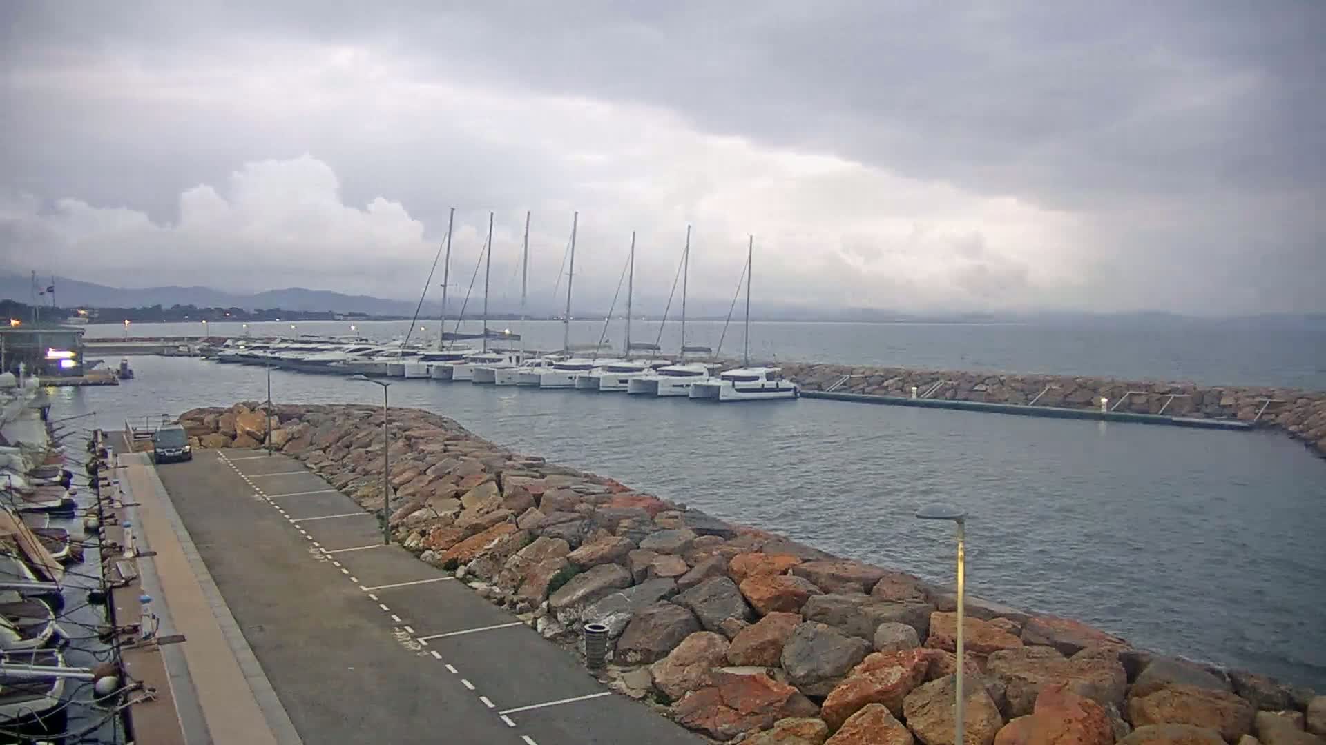 A marina with numerous sailboats moored, under a cloudy sky, is bordered by a rock wall and a paved area.