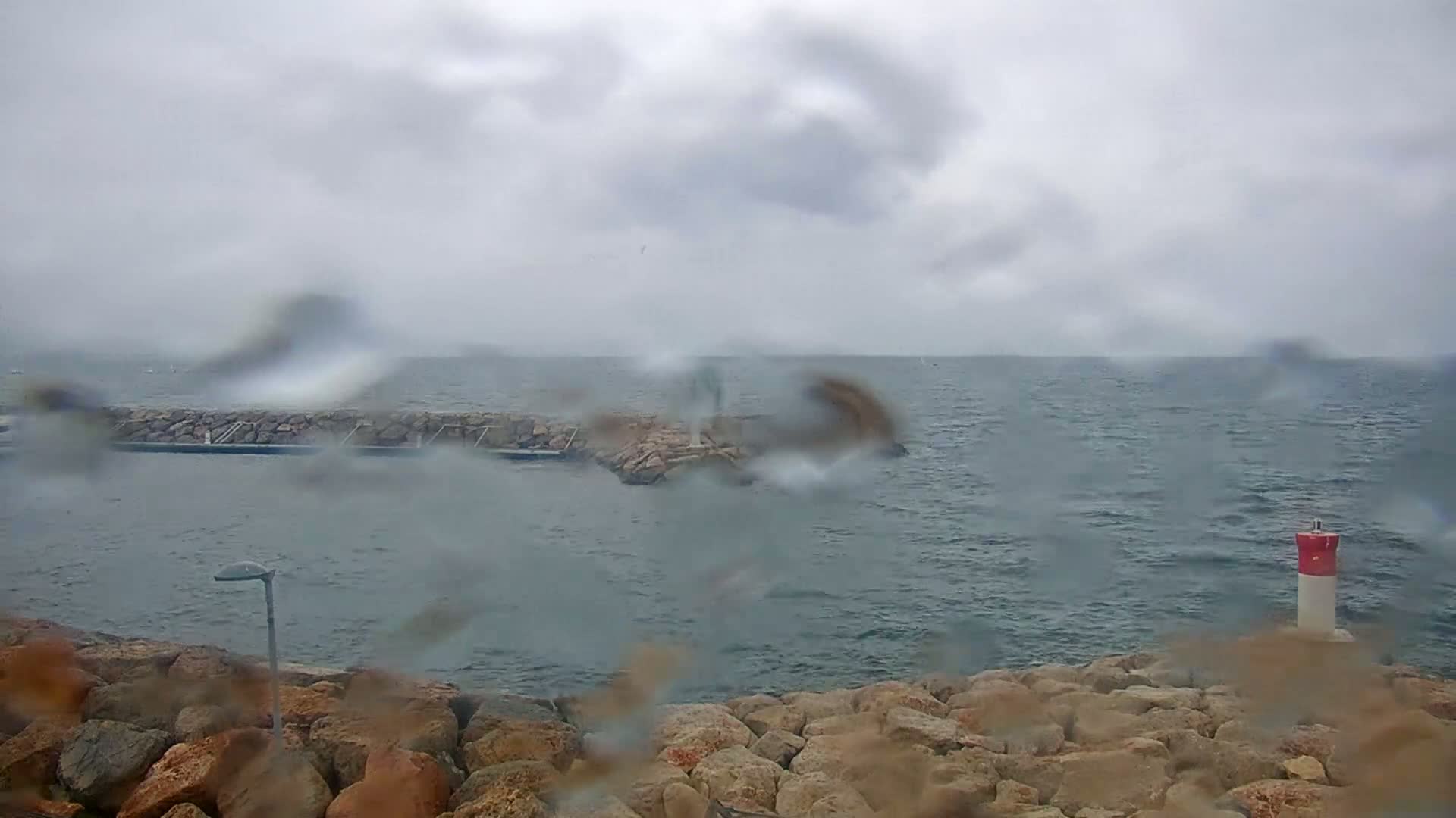 A rain-streaked view shows a rocky shoreline and a small breakwater in a choppy sea under a cloudy sky.