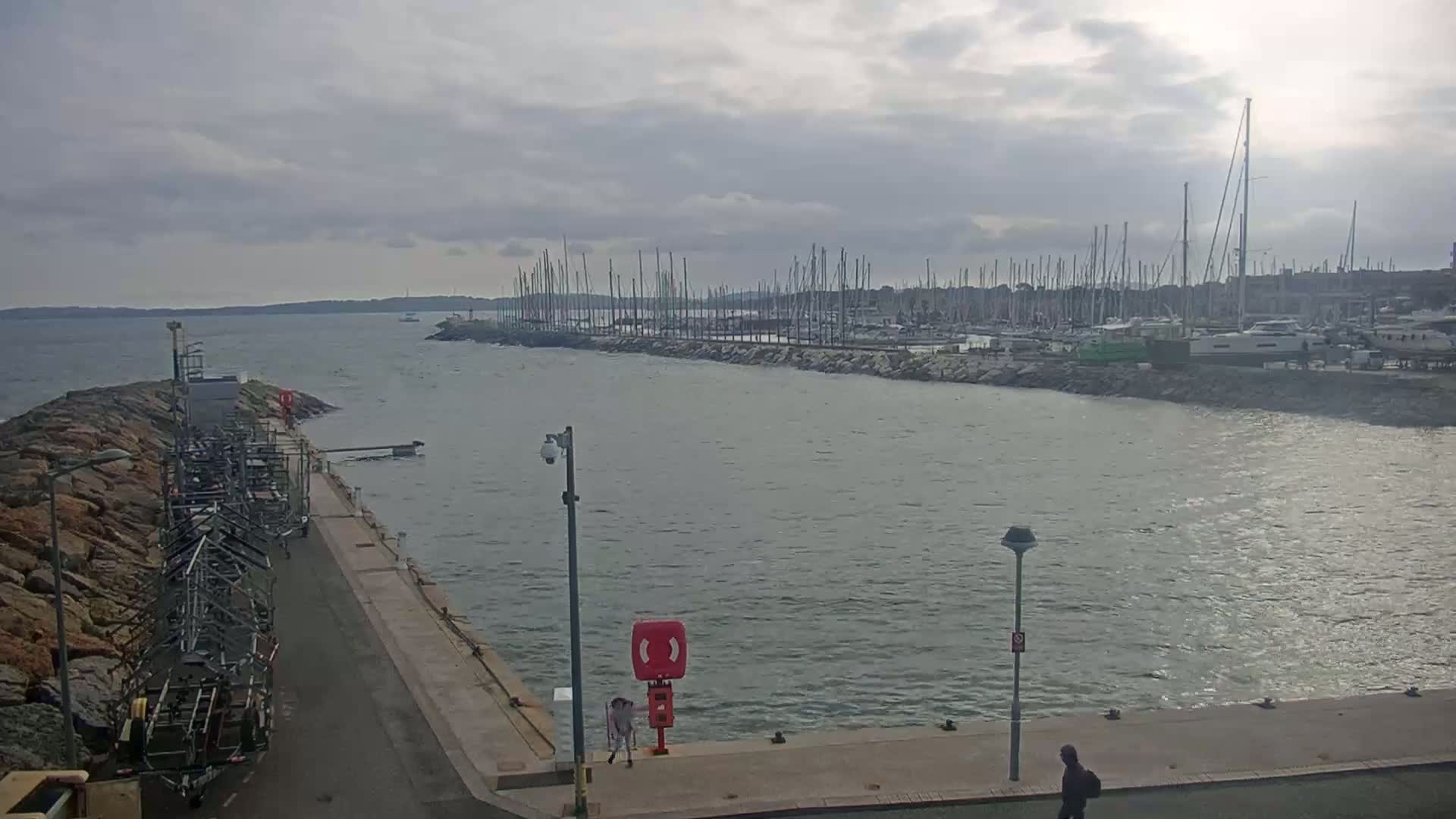 A harbor filled with numerous sailboats under a cloudy sky, with a calm sea and a person walking along the harbor wall.