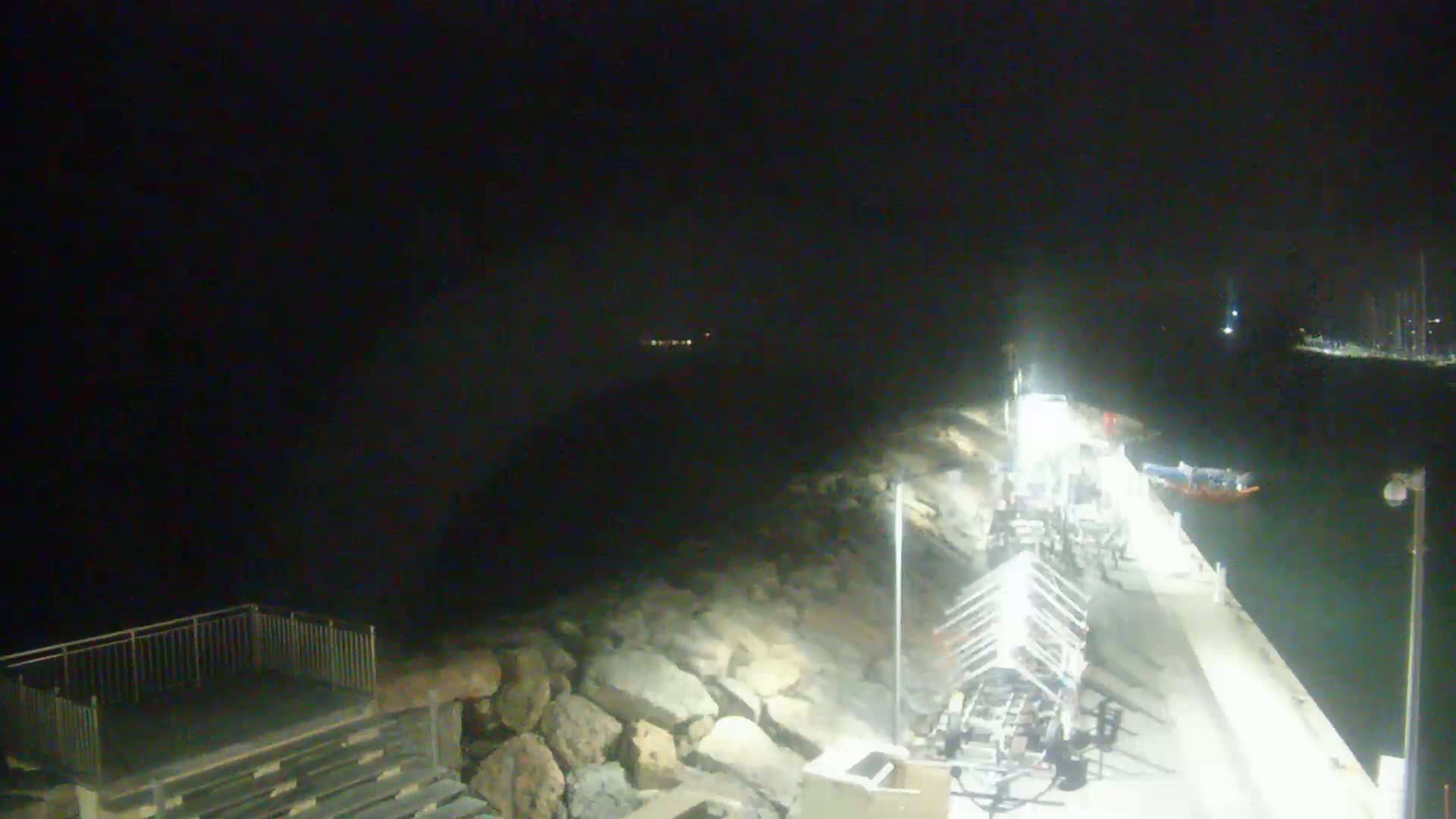 A nighttime view of a harbor with boats moored along a rocky embankment, illuminated by artificial light.