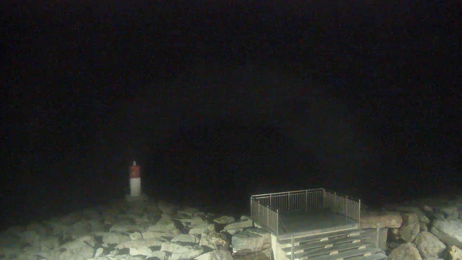 A small, dark-colored lighthouse stands on a rocky shoreline at night, next to a set of stairs and a metal railing.